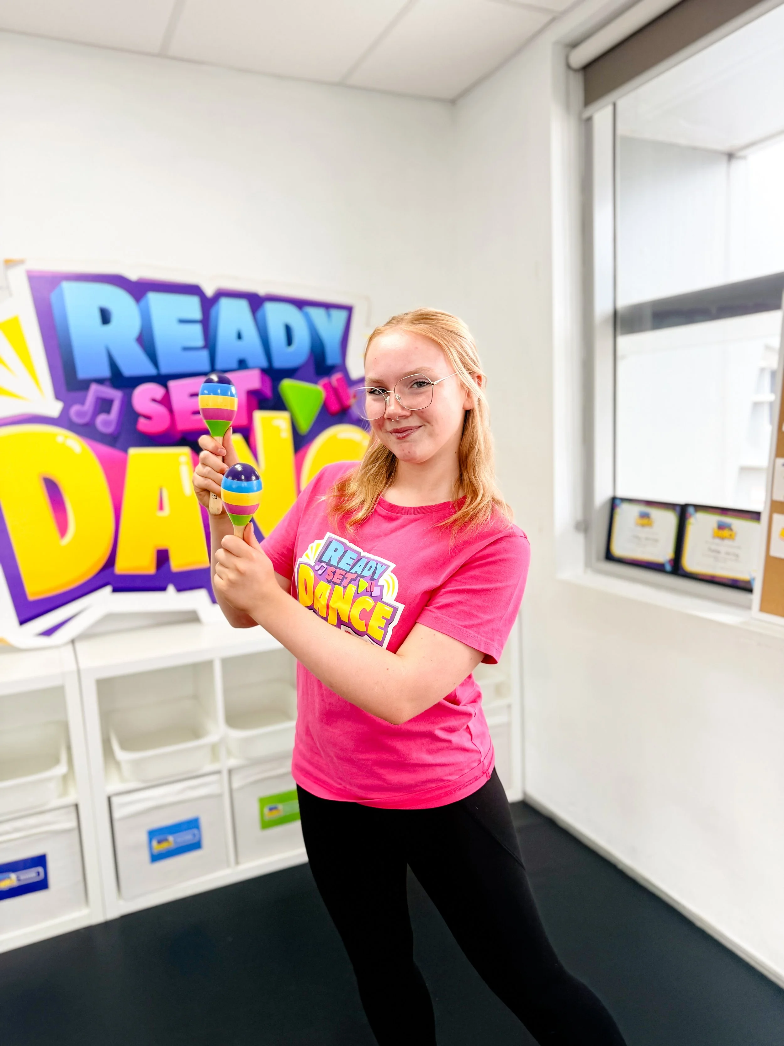 Teen girl in pink shirt holding colorful maracas, standing in front of a 'Ready Set Dance' sign, in a room with white walls and storage bins.