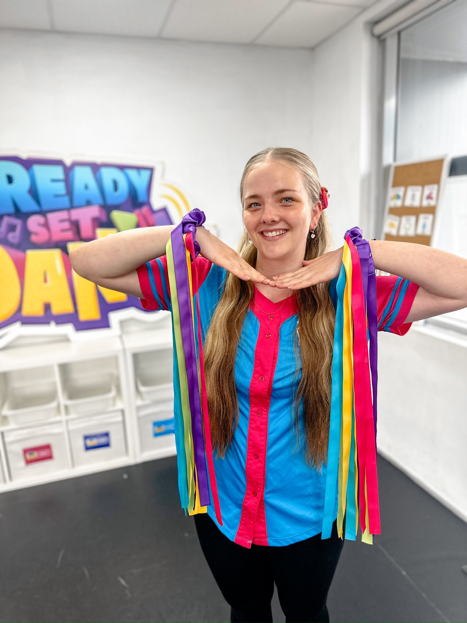 A young woman with long, light brown hair and a bright smile, wearing a colorful short-sleeved shirt and black pants, stands in a room decorated for a celebration, holding two multicolored ribbons around her shoulders, with a vibrant sign in the back