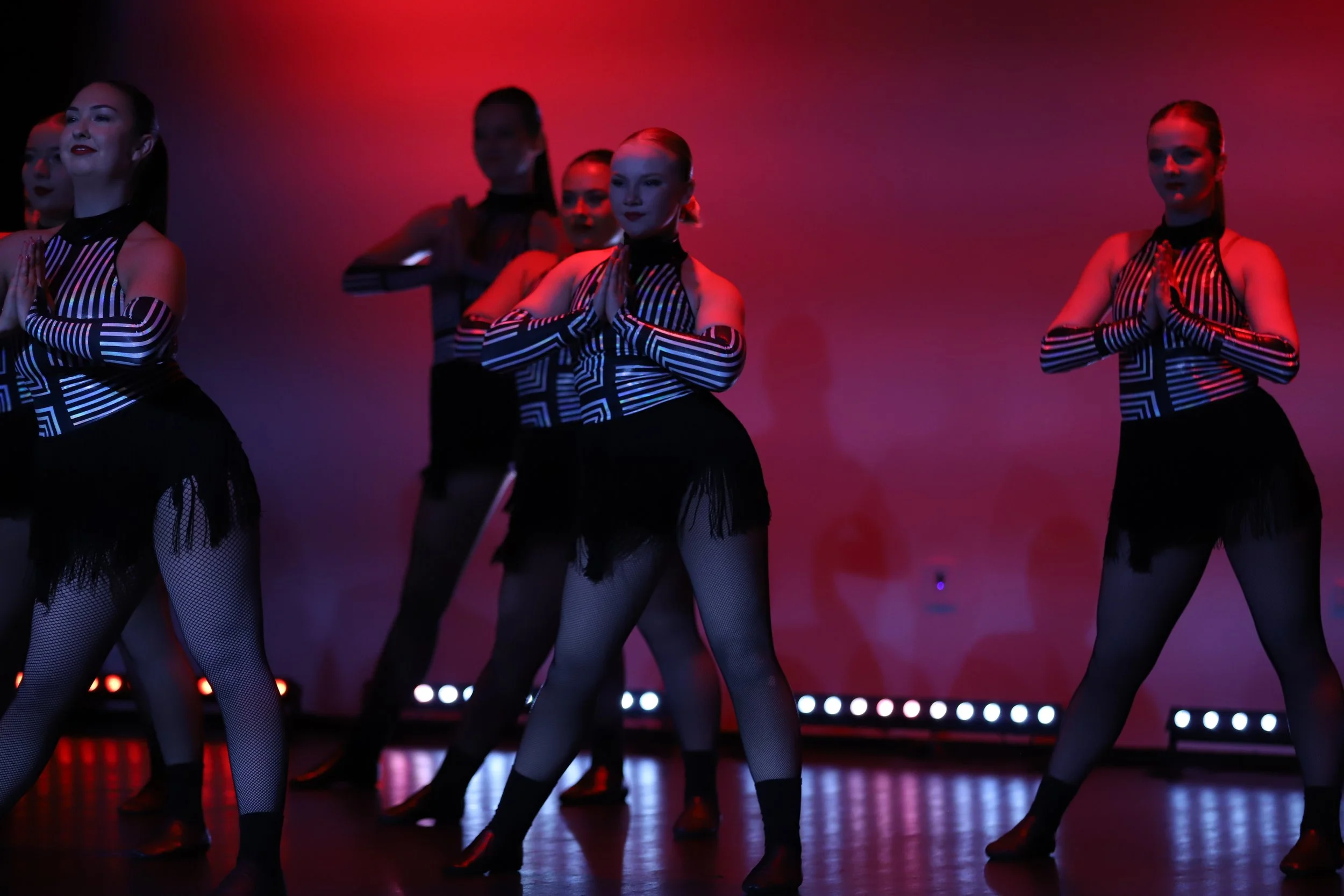 Group of women in black and white costumes performing a dance on stage with colorful lighting.