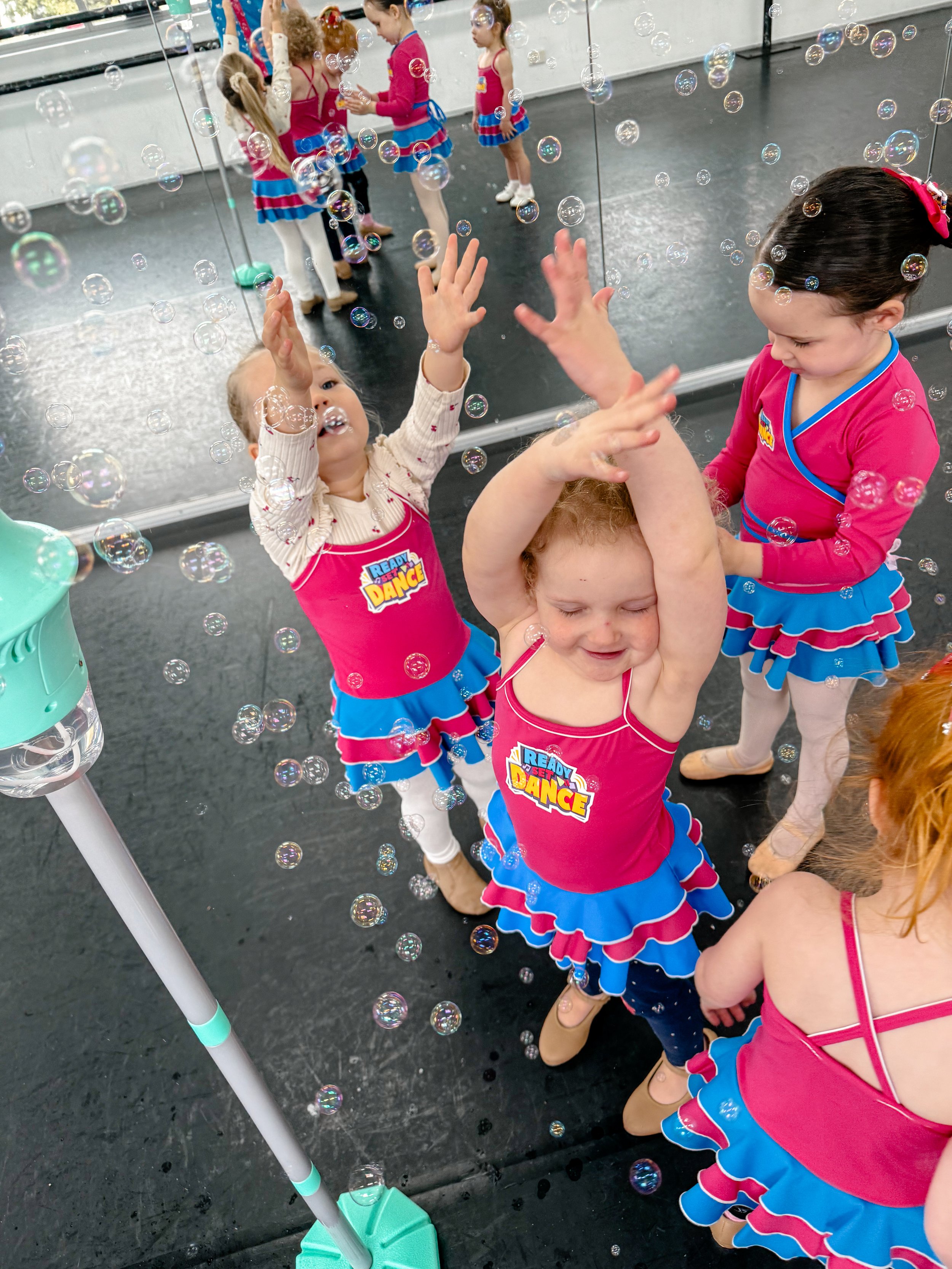 Young girls in colorful dance costumes playing with bubbles in a dance studio, with a mirrored wall reflecting their activities.