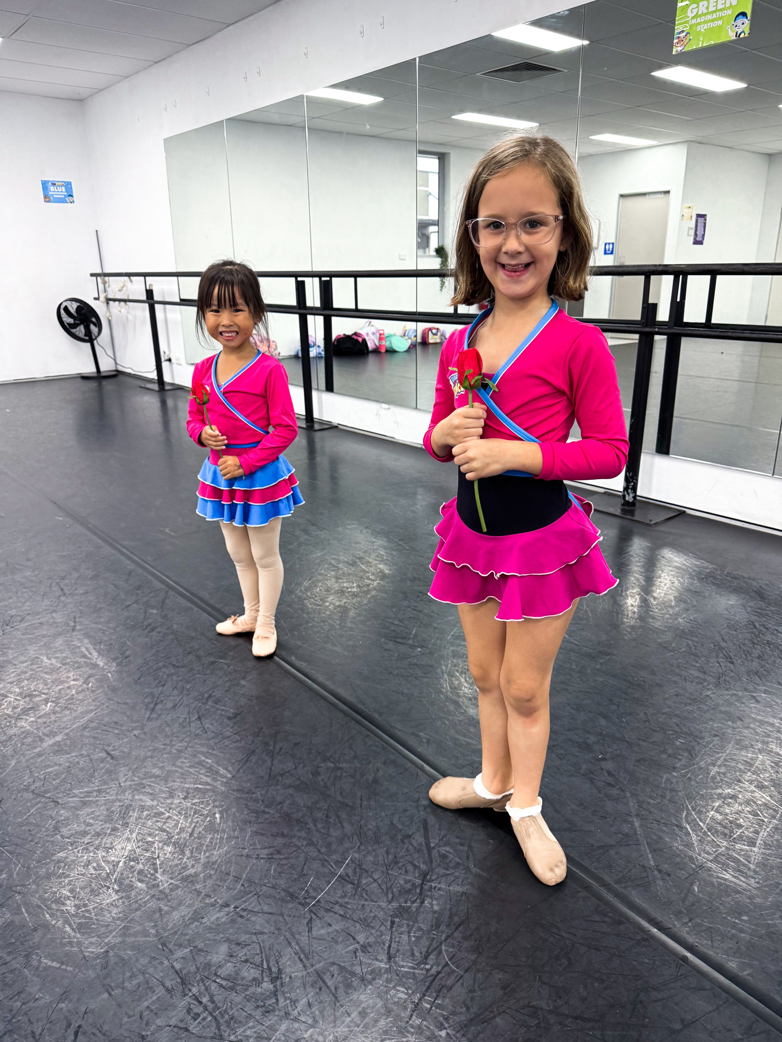 Two young girls in pink and blue ballet costumes standing in a dance studio, holding red roses, smiling at the camera.