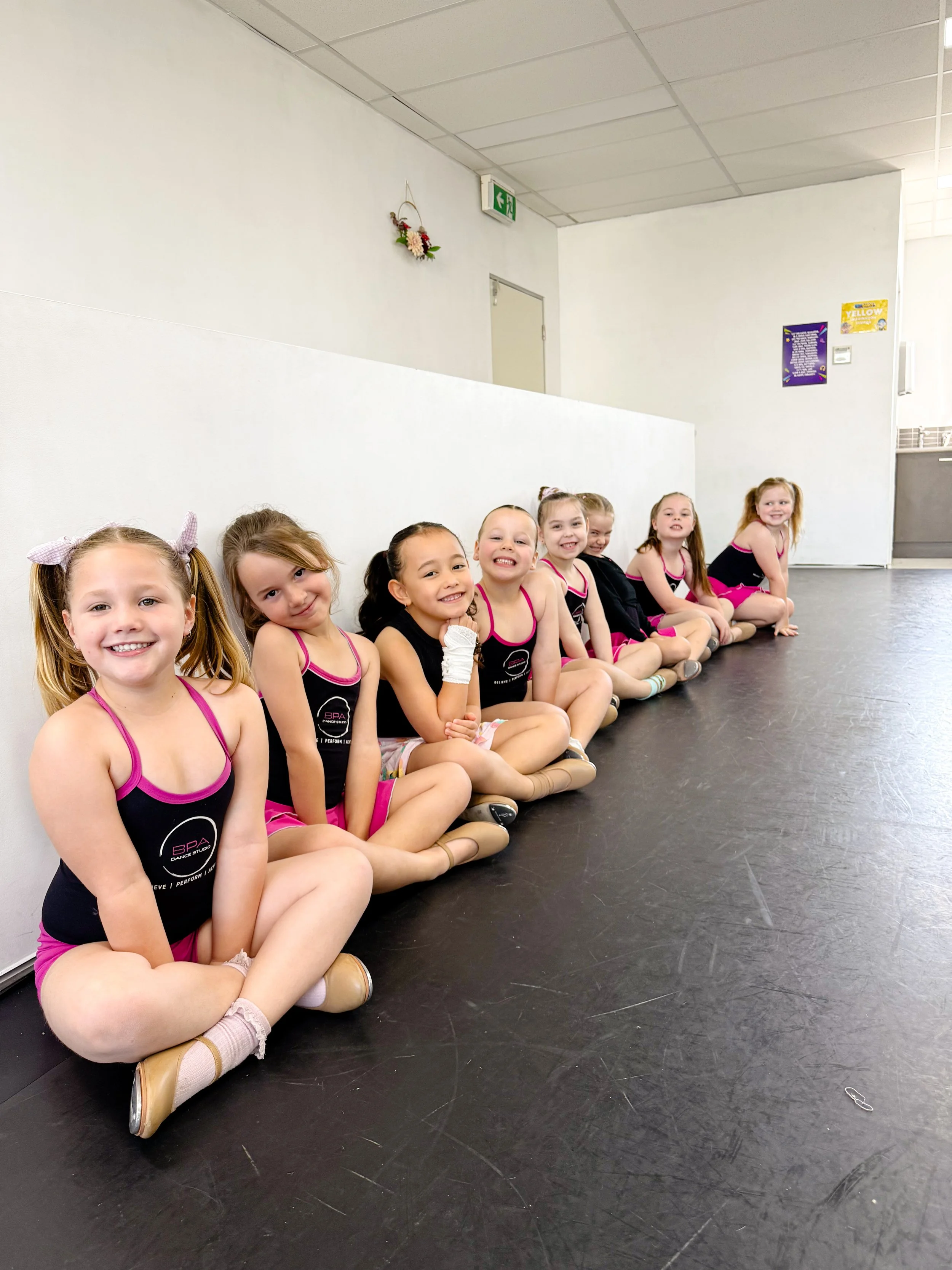 Group of young girls in matching dance outfits sitting on the floor against a white wall in a dance studio.