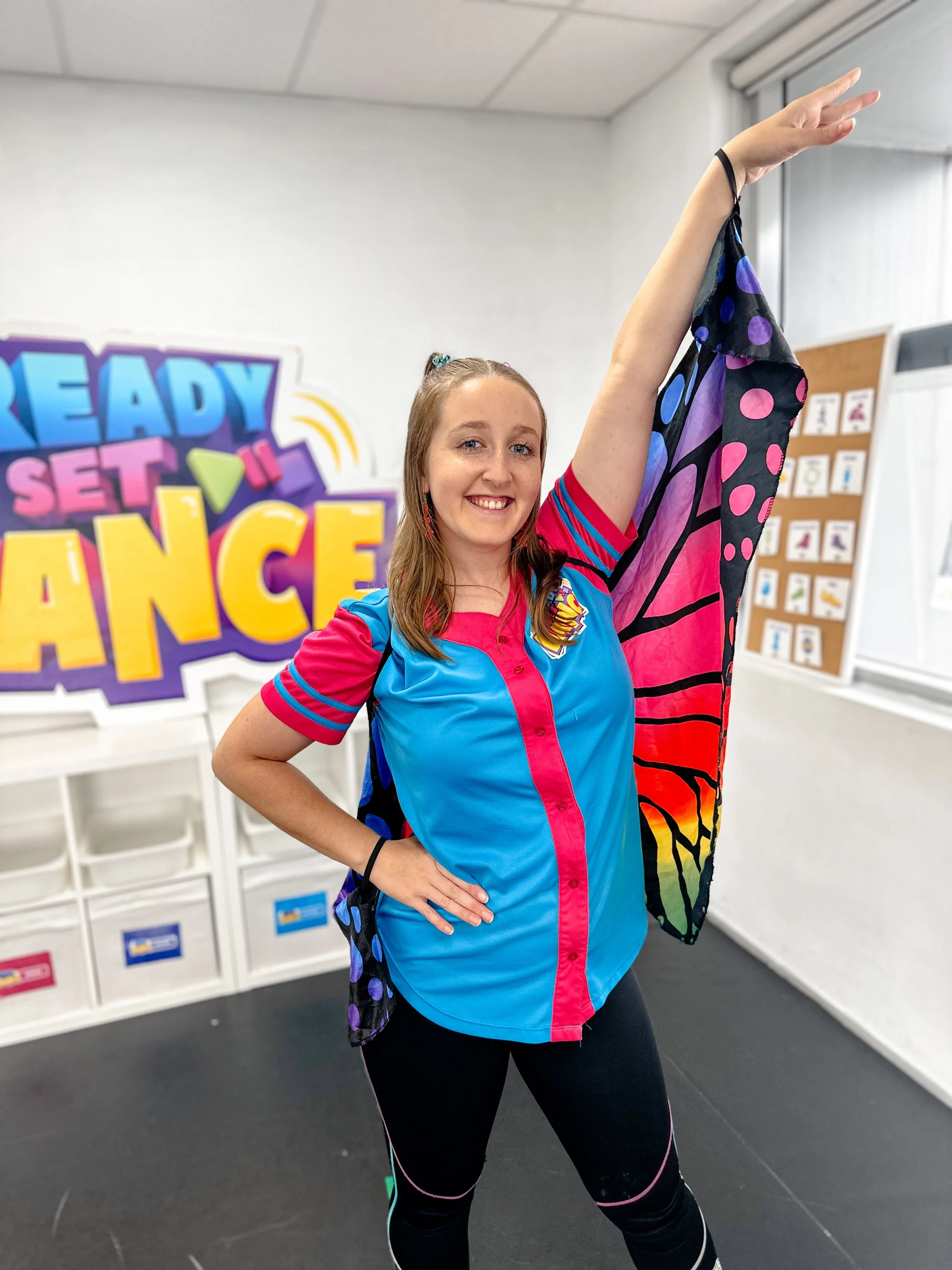 Young woman smiling and stretching in a dance pose at a dance studio with a colorful 'Ready Set Dance' sign in the background.
