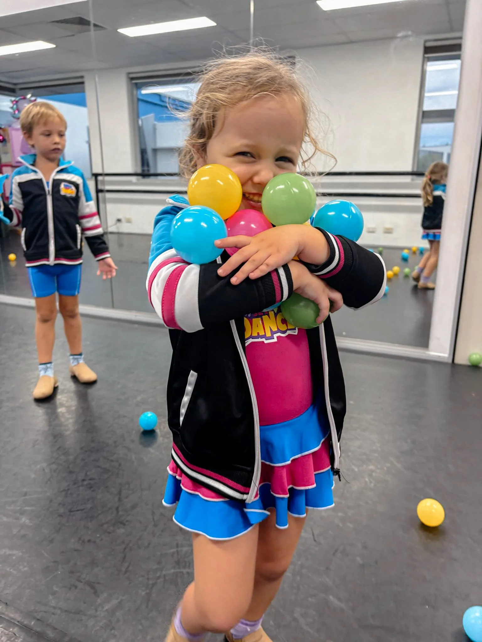 A young girl in a colorful cheerleader uniform holding multiple plastic balls, smiling at the camera, in an indoor play area or gym with other children and scattered balls in the background.