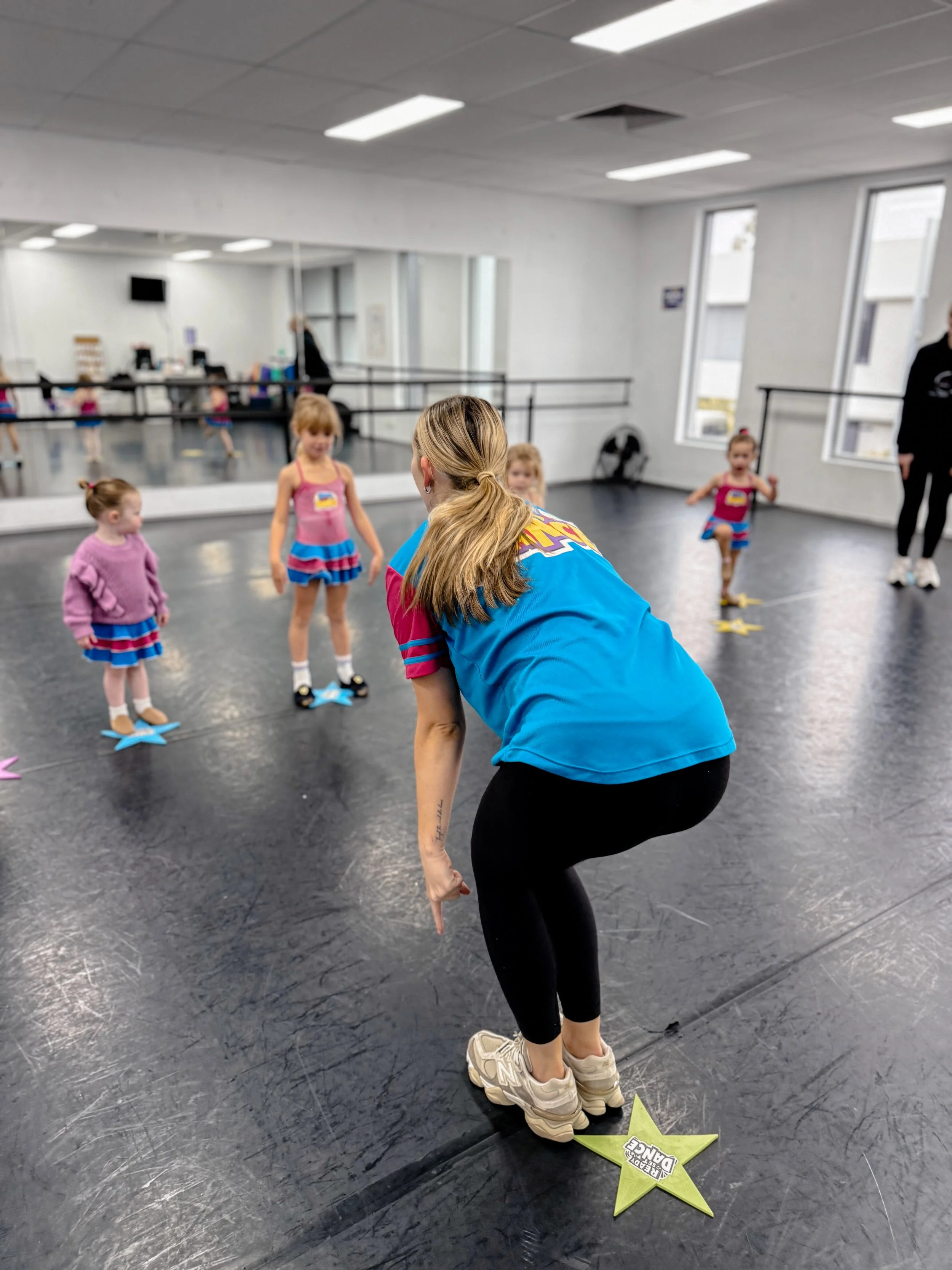 A dance instructor or teacher is demonstrating a move to young children in a dance studio with a black floor, mirrored wall, and large windows. The children are dressed in colorful dance outfits, and the instructor is bent over slightly with one leg 