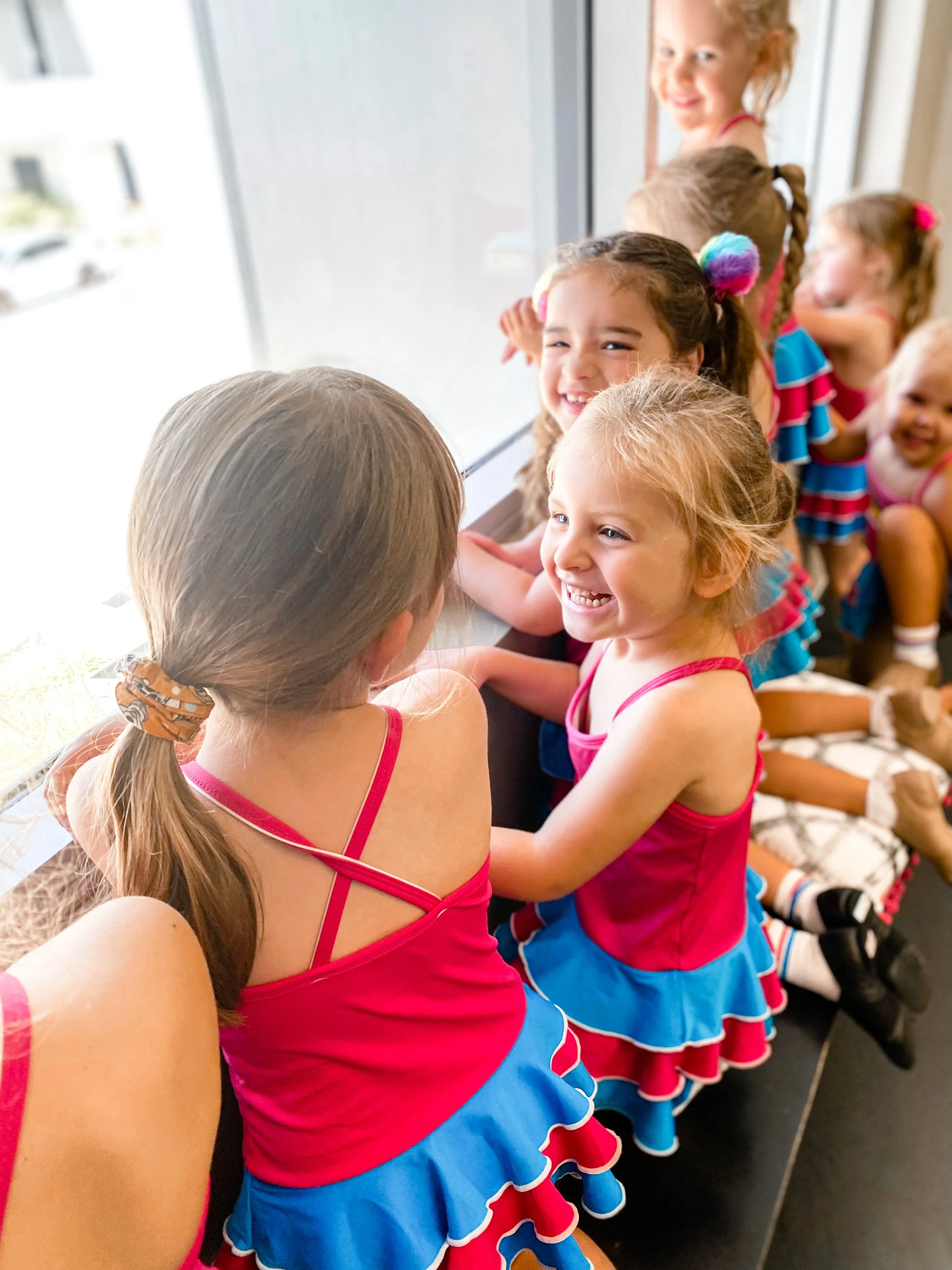 Group of young girls dressed in colorful cheerleading outfits sitting by a large window, smiling and chatting.