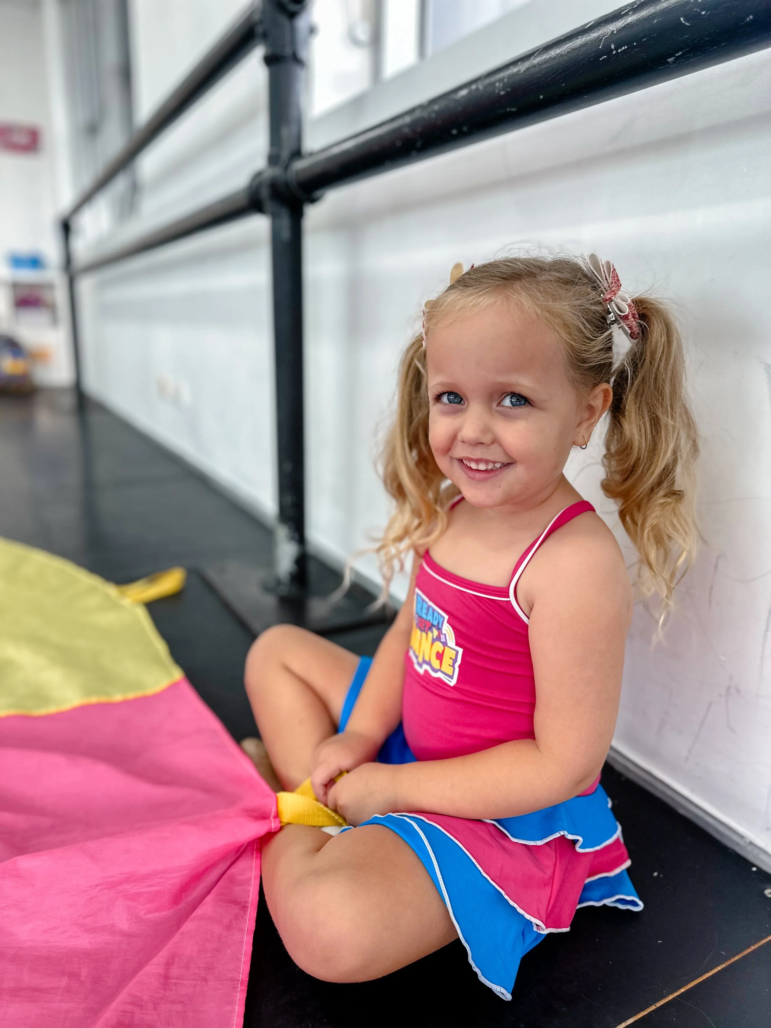 A young girl with blonde hair in pigtails, wearing a pink and blue cheerleading outfit, sitting on the floor and smiling at the camera, holding a yellow and pink portion of a colorful parachute or fabric.