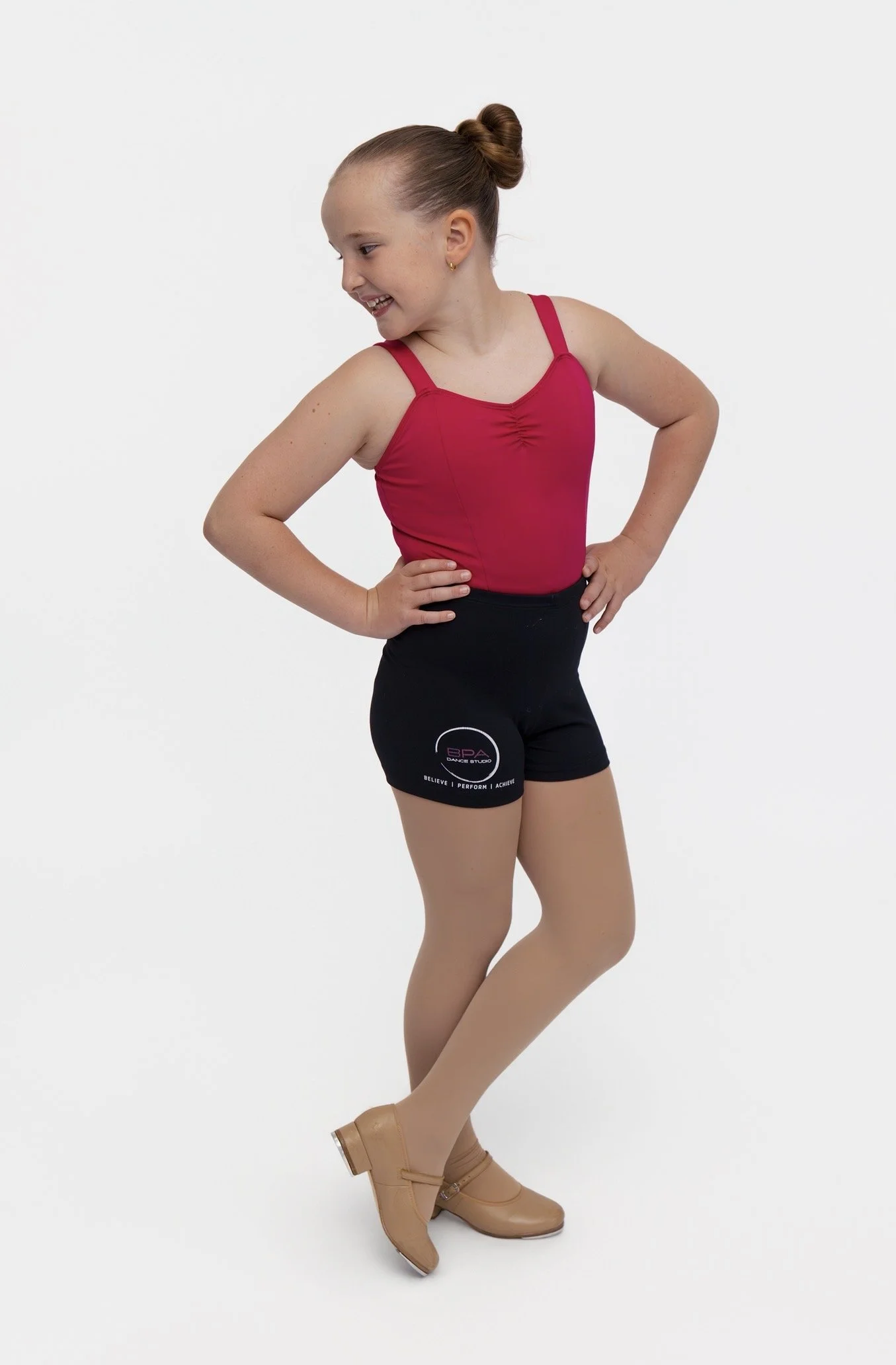 Young girl with a bun hairstyle wearing a pink dance leotard, black shorts with a logo, tan tights, and beige dance boots, posing with hands on hips against a plain white background.
