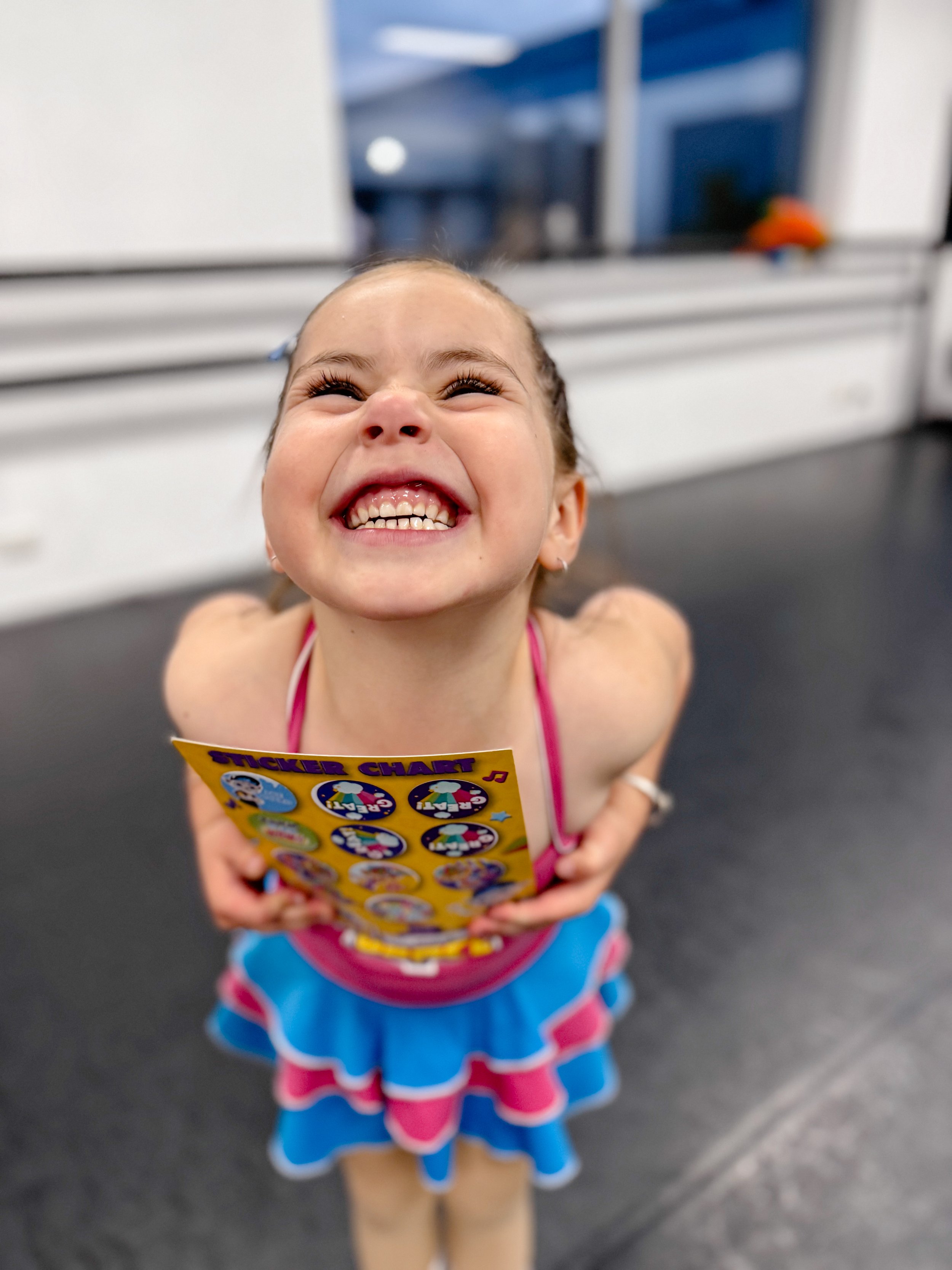Young girl with a big smile, showing teeth, holding a sticker chart, wearing a colorful skirt and pink tank top, standing indoors.