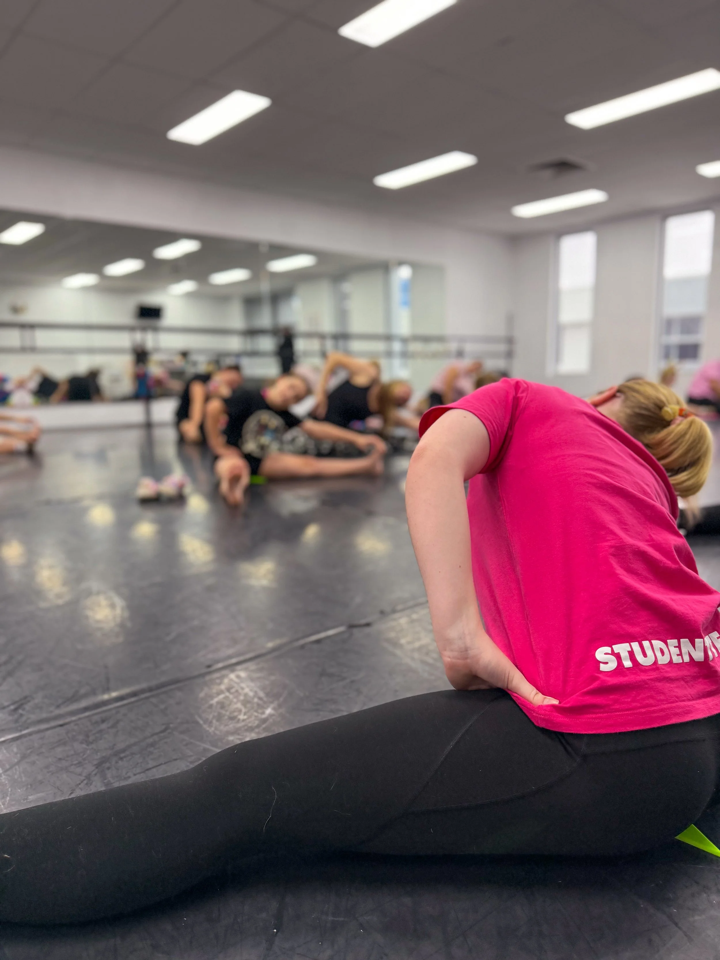 A fitness class in a dance studio with several women stretching on the floor, one woman in the foreground wearing a pink shirt and black leggings stretching her back while sitting with one leg extended and the other bent. The studio has a large mirro