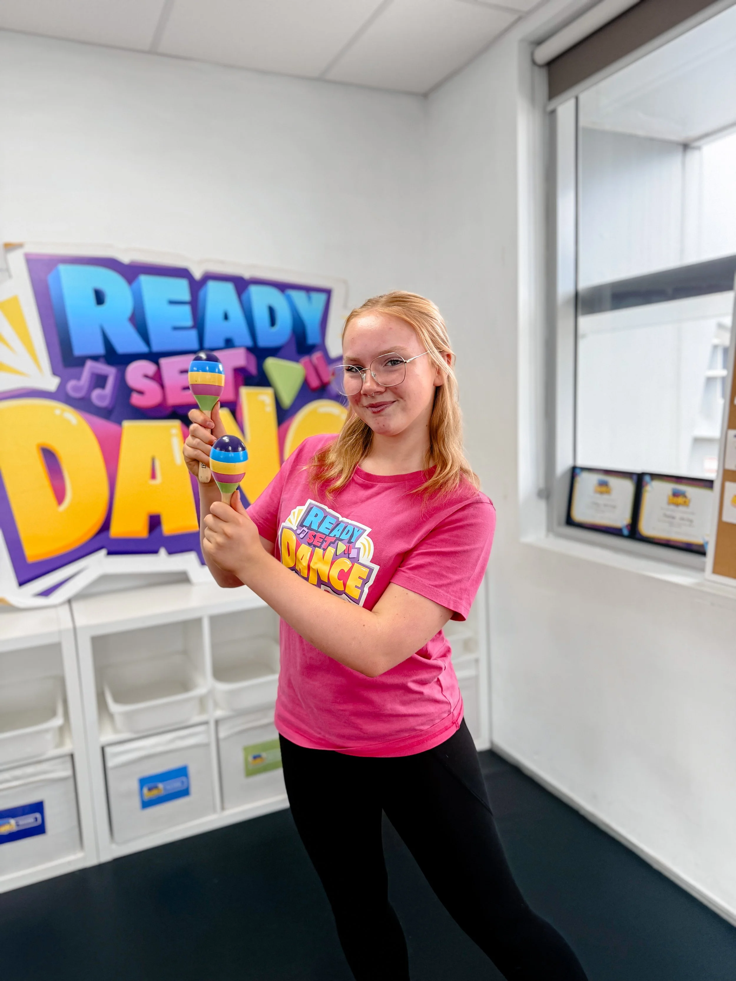 Young woman in pink shirt holding maracas in a dance studio with vibrant 'Ready Set Dance' sign in the background.
