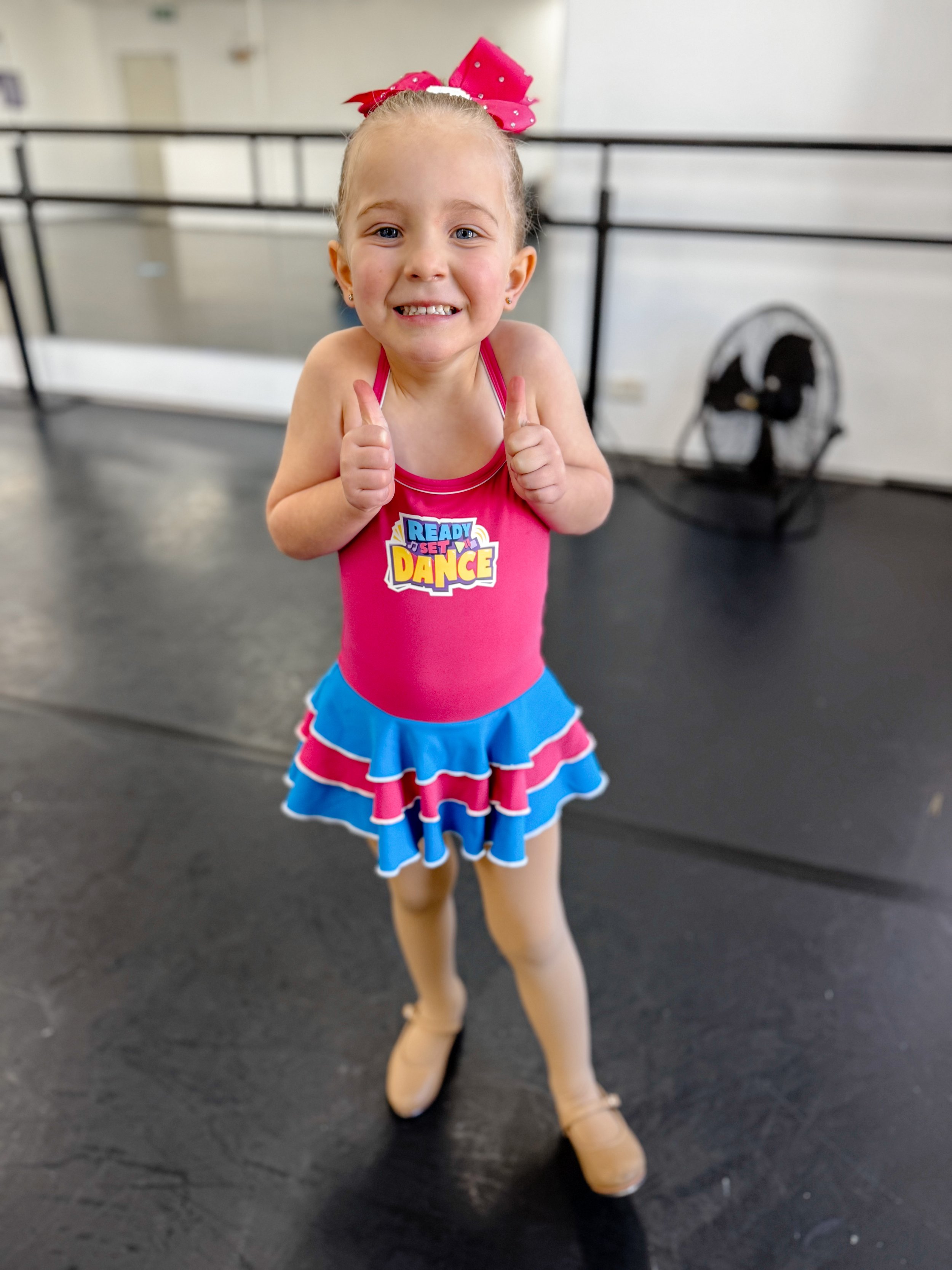 Young girl in vibrant pink and blue cheerleading outfit smiling and giving two thumbs up inside a dance studio.