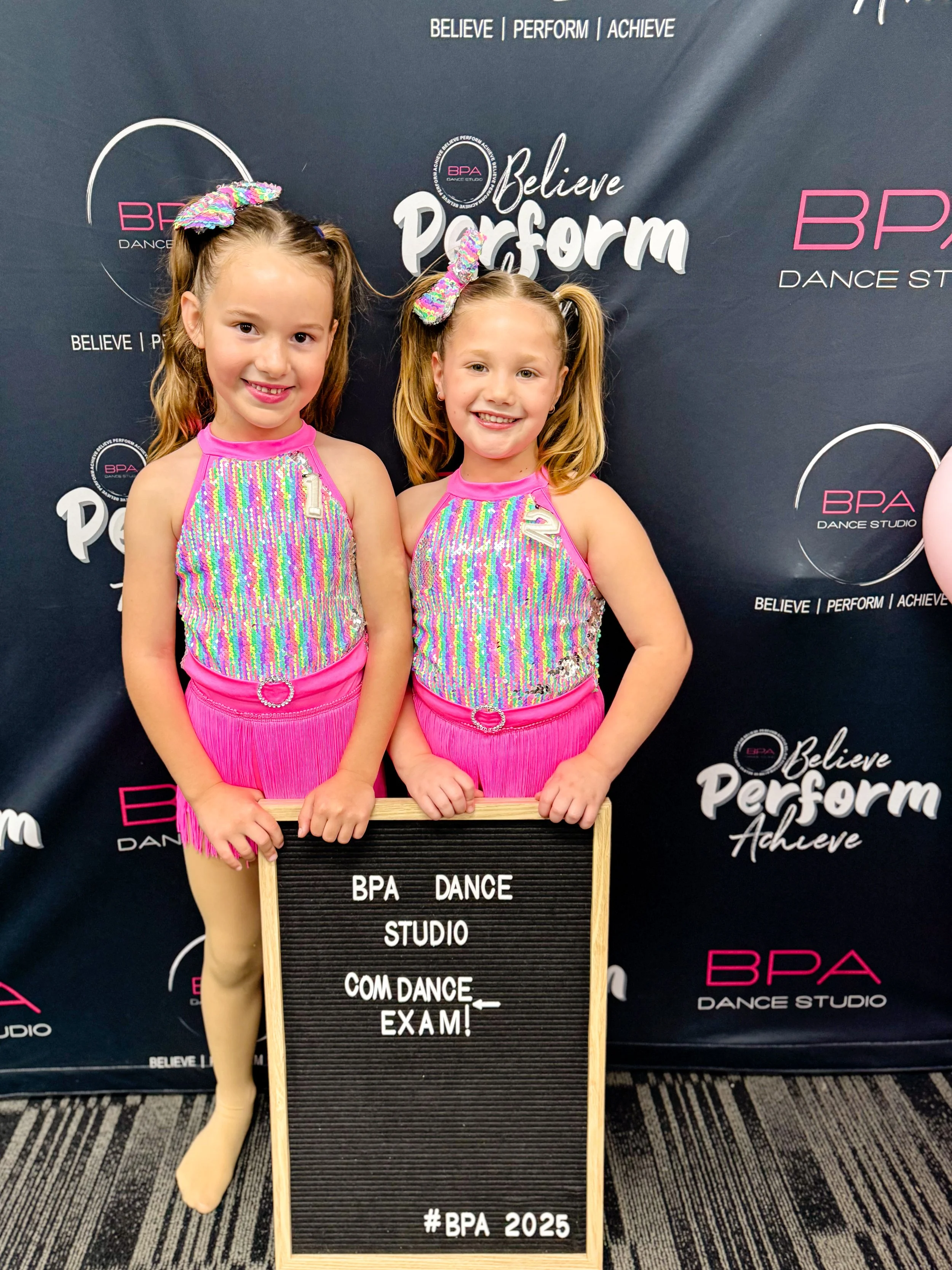 Two young girls in colorful dance costumes are posing together, holding a black letter board with white text at a dance studio event. The background features a dark banner with the words 'Believe Perform Achieve' and 'BPA Dance Studio' printed on it.