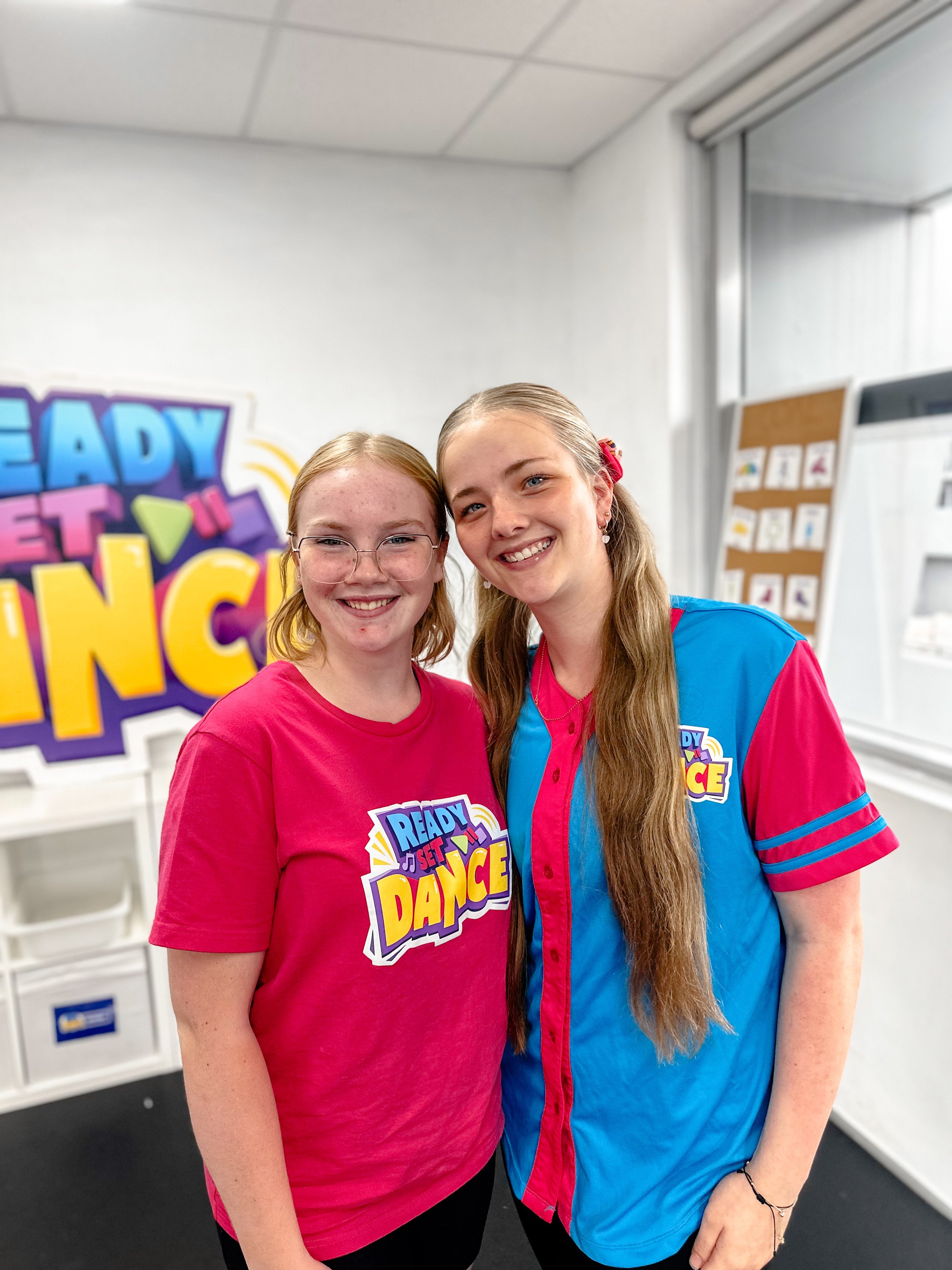 Two young girls smiling at the camera in a classroom with a large colorful sign that says 'Ready Set Dance' in the background.