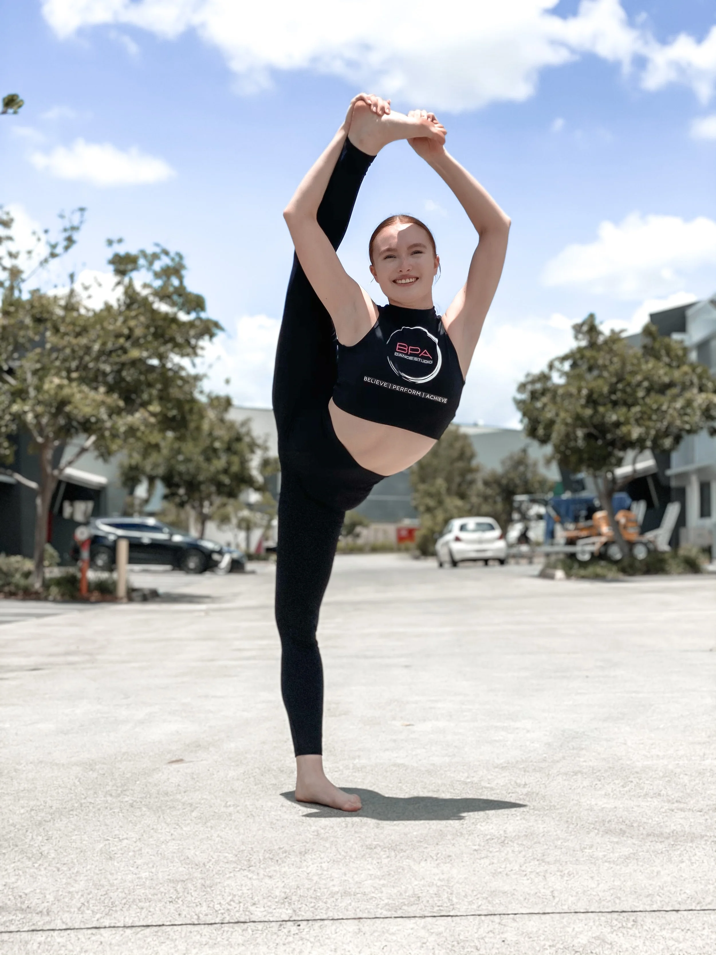 A young woman practicing yoga outdoors on a sunny day, performing a standing bow yoga pose with one leg lifted, holding her foot behind her, and smiling.