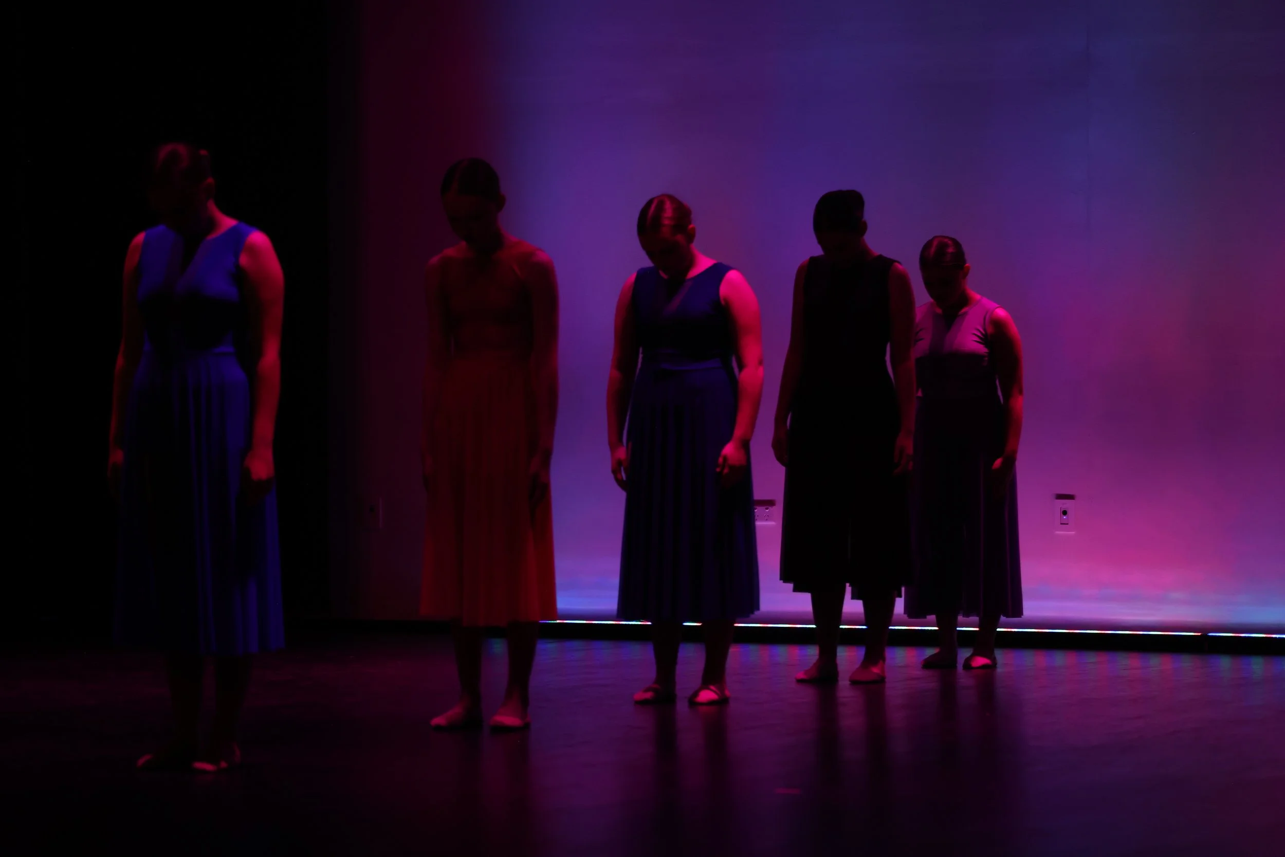 Five women standing in a row on a stage with purple and pink lighting, facing downward, wearing long dresses.