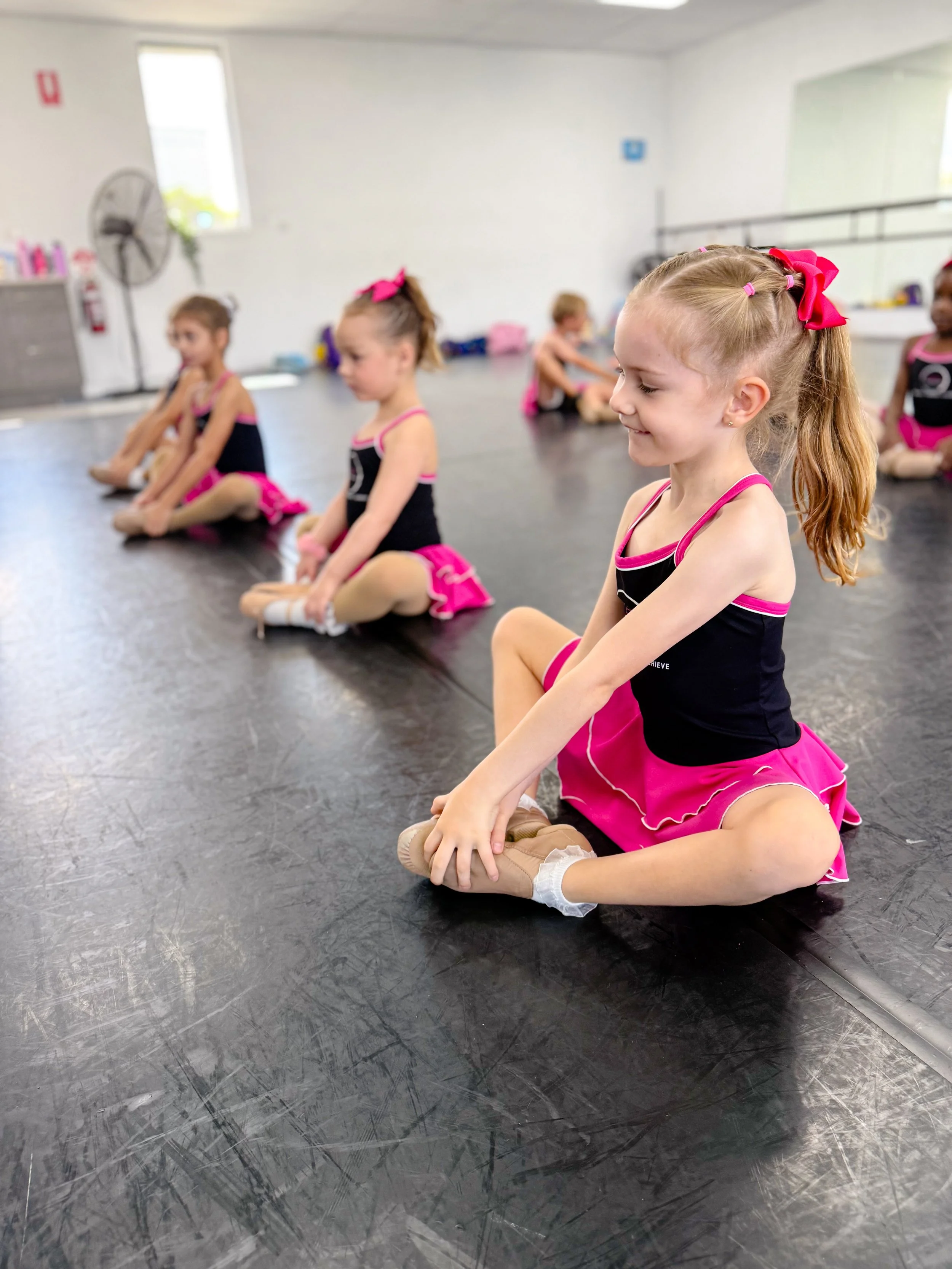 Young girls in ballet costumes sitting on the floor during a dance class in a studio.