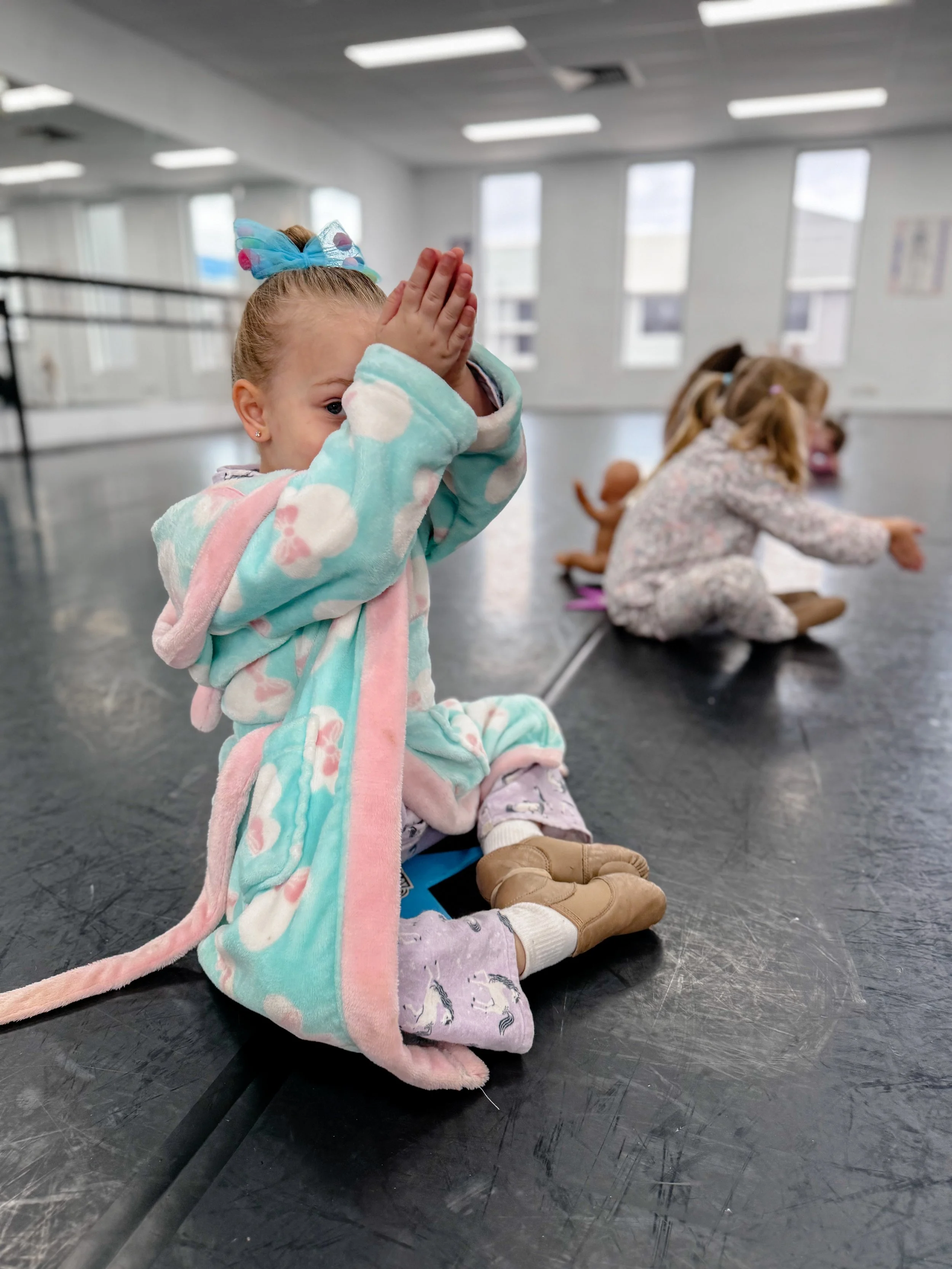A young girl sitting on the floor with her knees bent, covering her face with her hands. She is wearing pajamas, a colorful robe, and a blue bow in her hair, in a dance studio with big windows and other children in the background.