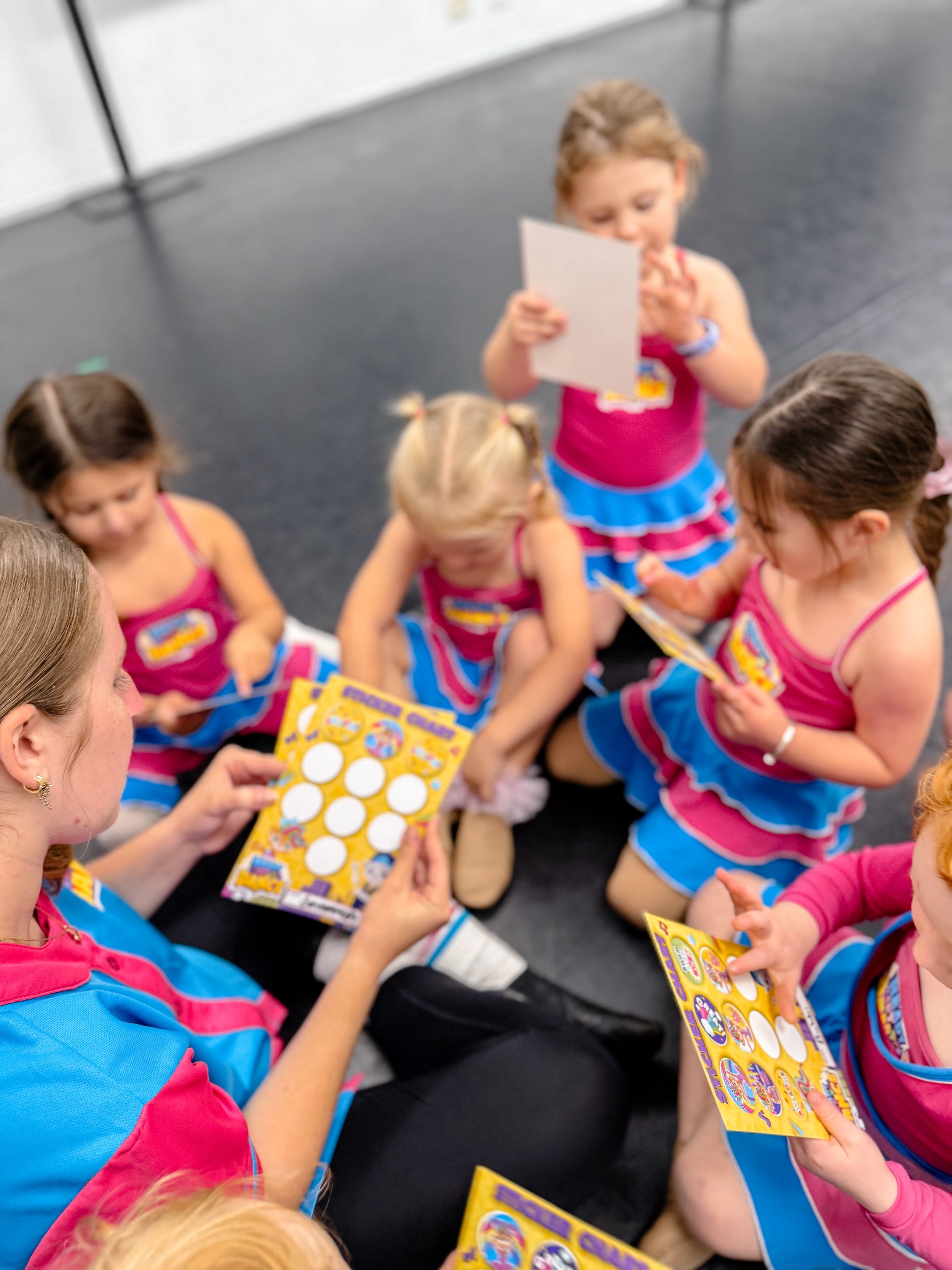 Young girls sitting on the floor, wearing cheerleading uniforms, while looking at bingo game cards, participating in a game or activity.