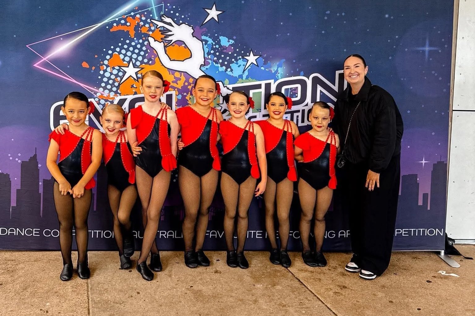 Group of seven young girls in black and red dance costumes with fishnet tights, standing with their dance instructor in front of a colorful backdrop for a dance competition.