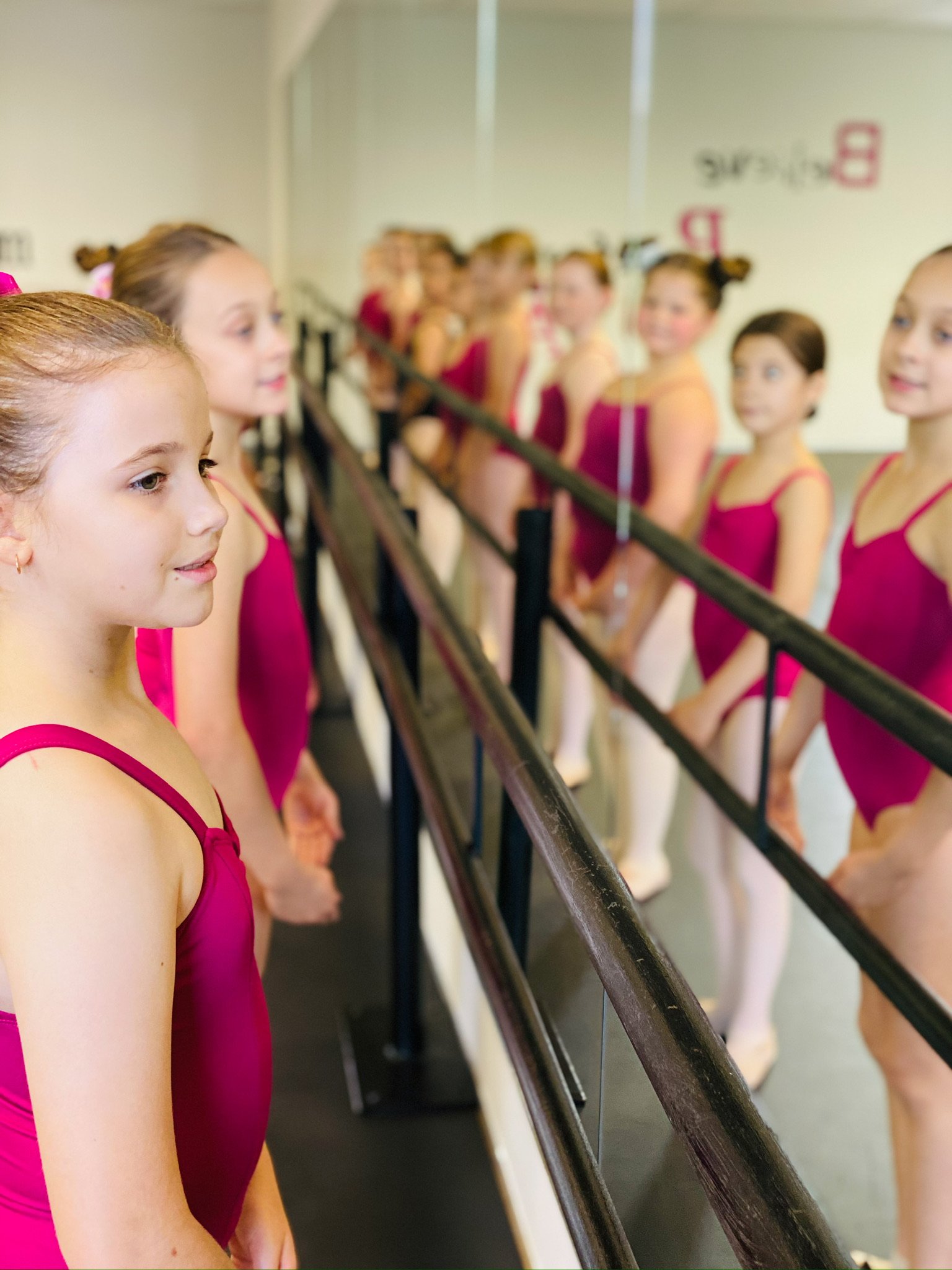Young girls in pink ballet leotards standing in front of a mirror, preparing for a ballet class at a dance studio.