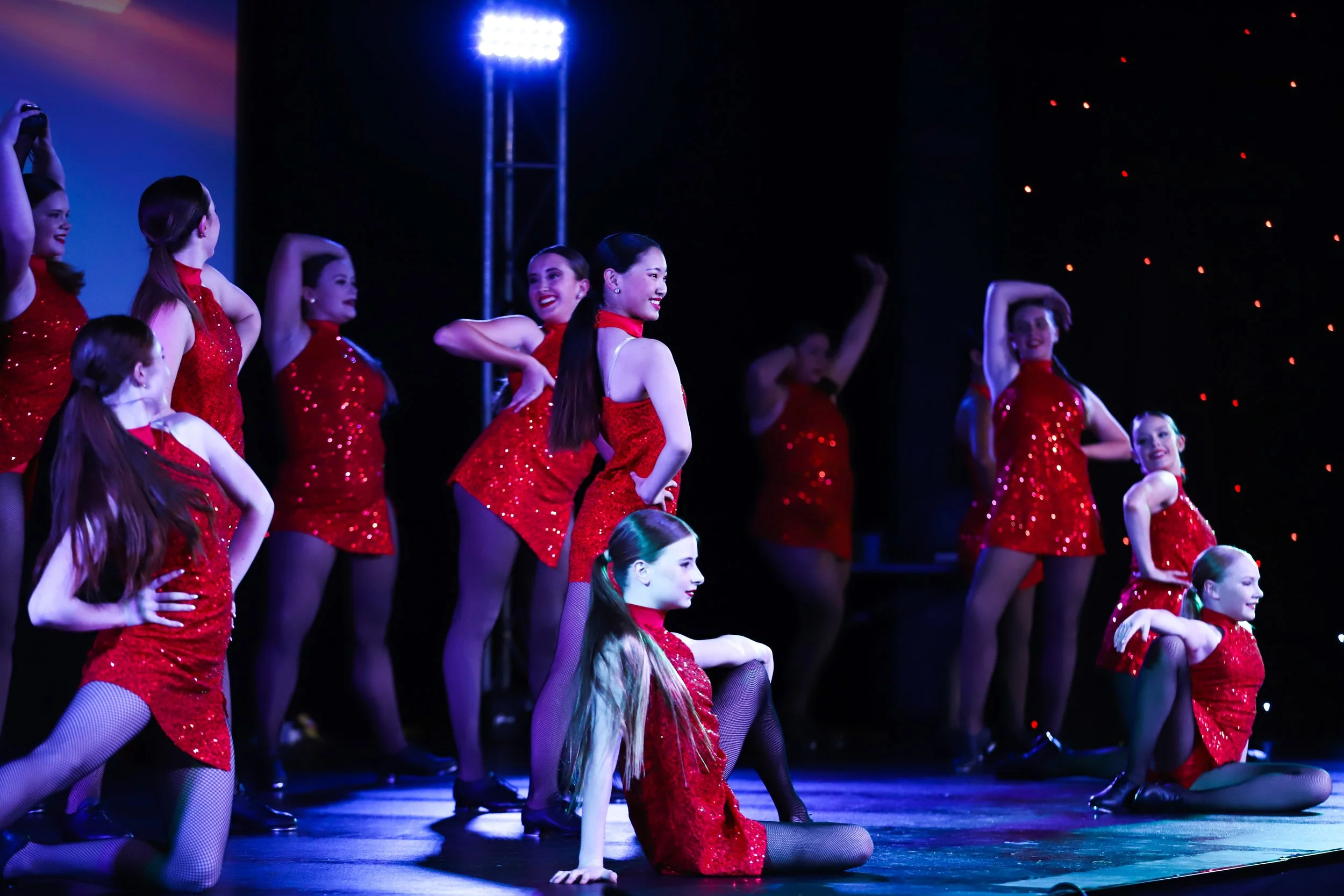 Group of young female dancers in red sequined dresses performing on stage with stage lights and a dark background.