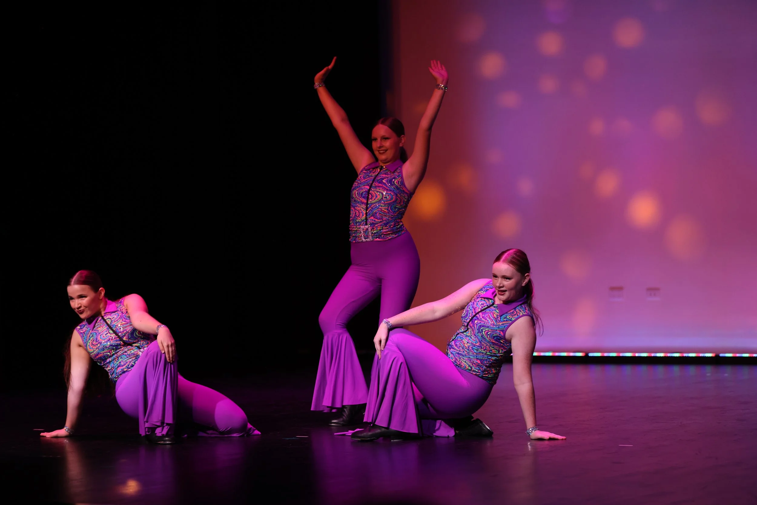 Three young women in colorful, matching dance costumes performing on stage with purple flooring and a pink and purple background. The women are in dynamic dance poses, smiling.