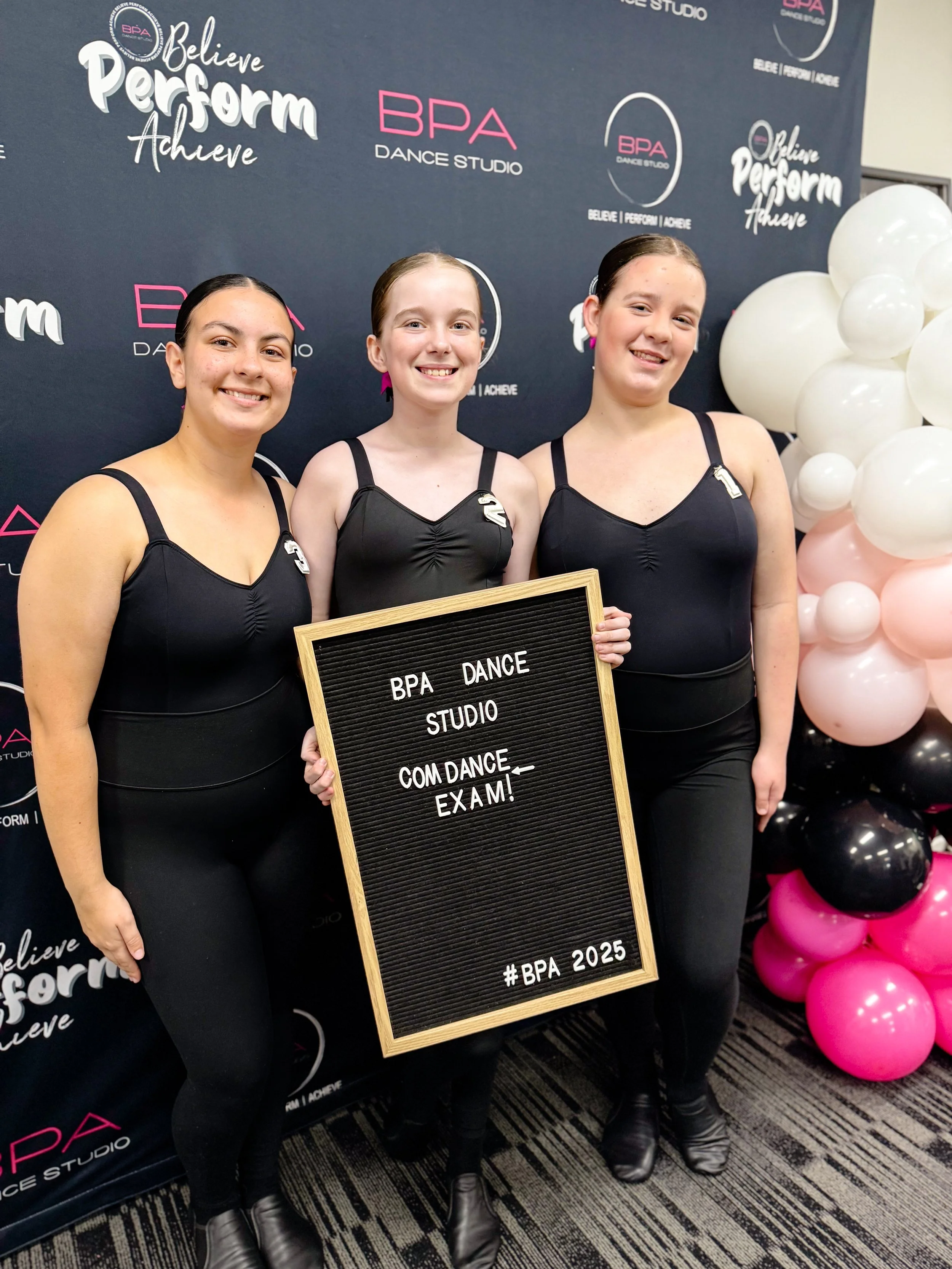 Three young women in black dance attire standing together smiling, holding a black letter board with 'BPA DANCE STUDIO COM DANCE EXAM! #BPA 2025', in front of a backdrop with the BPA Dance Studio logo and motivational phrases, next to pink, black, and white balloons.