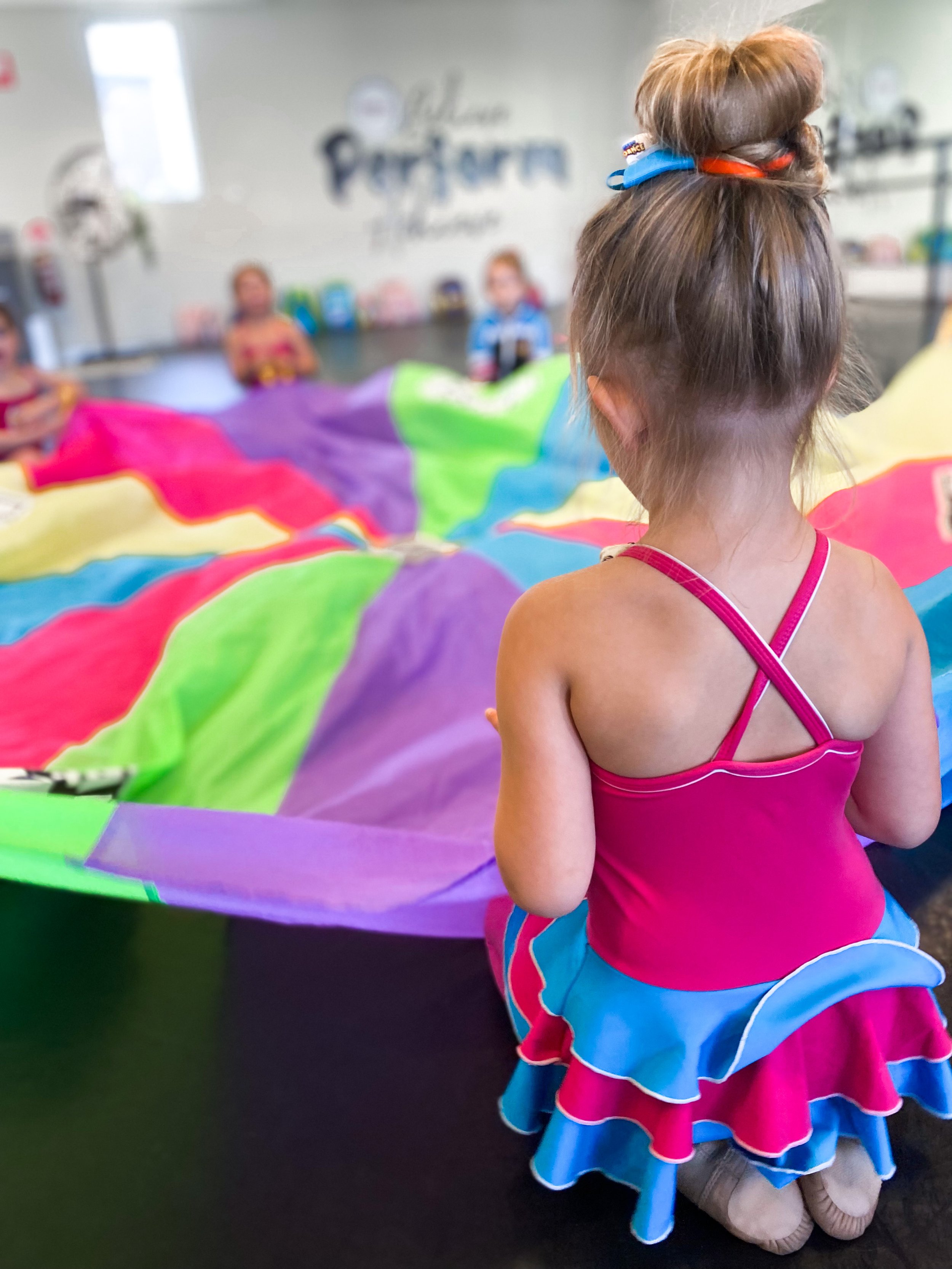 A young girl in a pink and blue dress standing in front of a group of children playing with a colorful parachute in a playroom.
