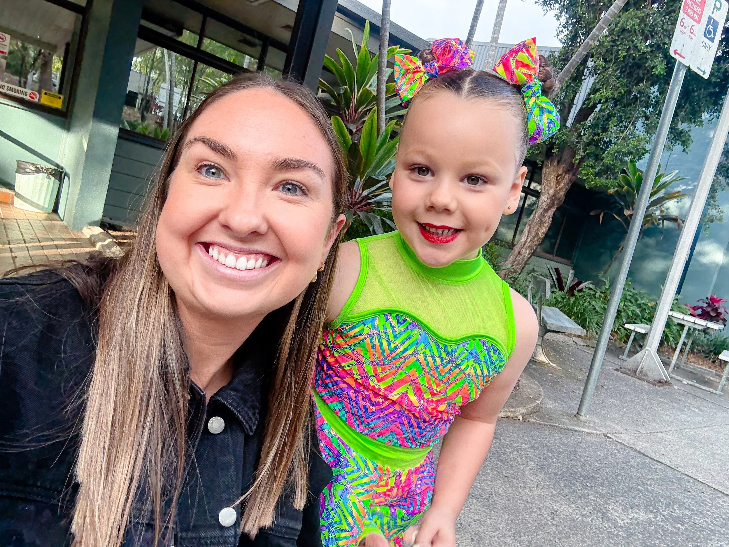 A woman and a young girl taking a selfie outdoors, both smiling at the camera. The girl is dressed in a colorful, neon green and patterned costume with a large, rainbow-colored bow on her head.