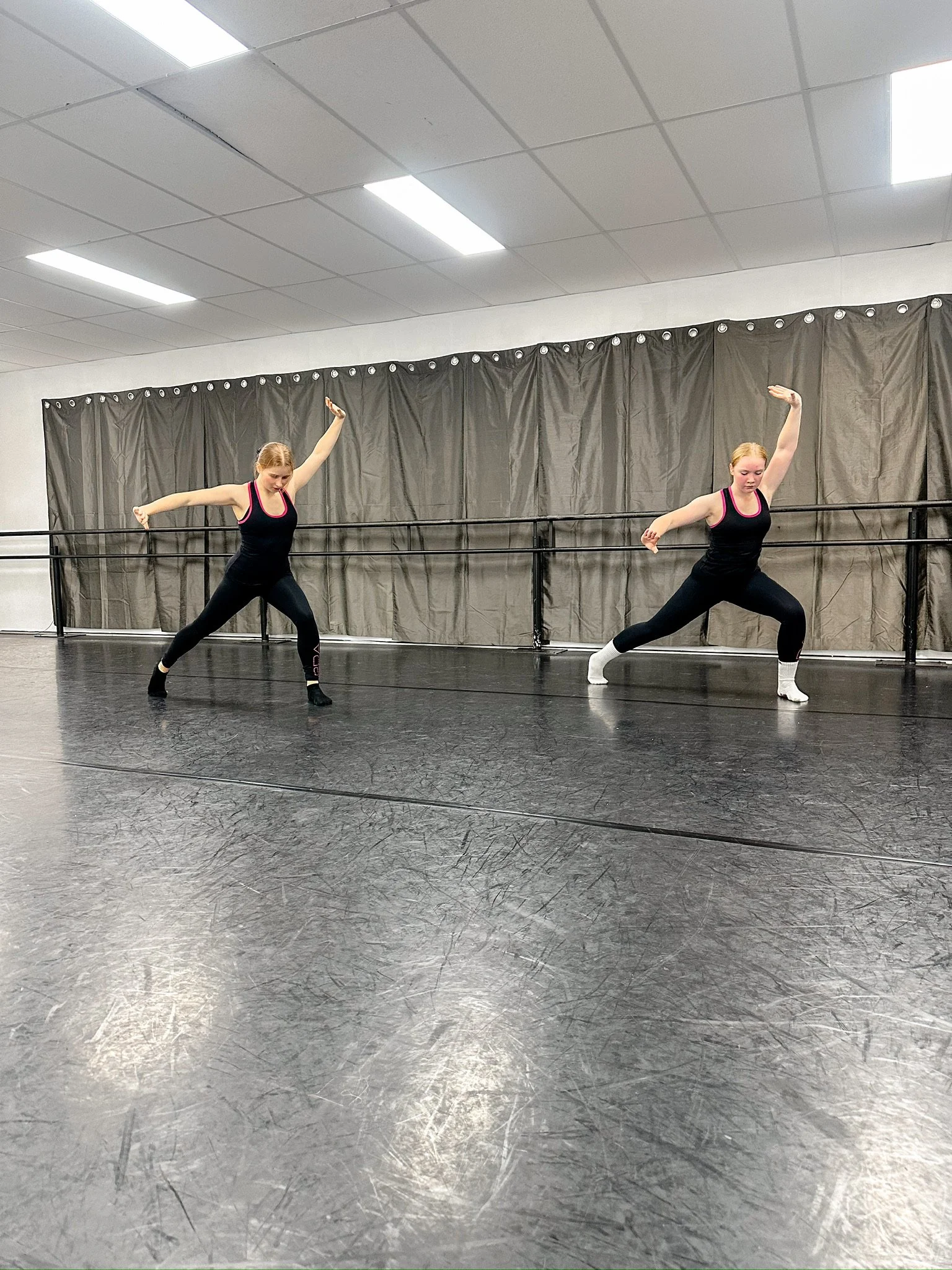 Two young women practicing dance moves in a dance studio with black flooring, black curtains, and white ceiling lighting.