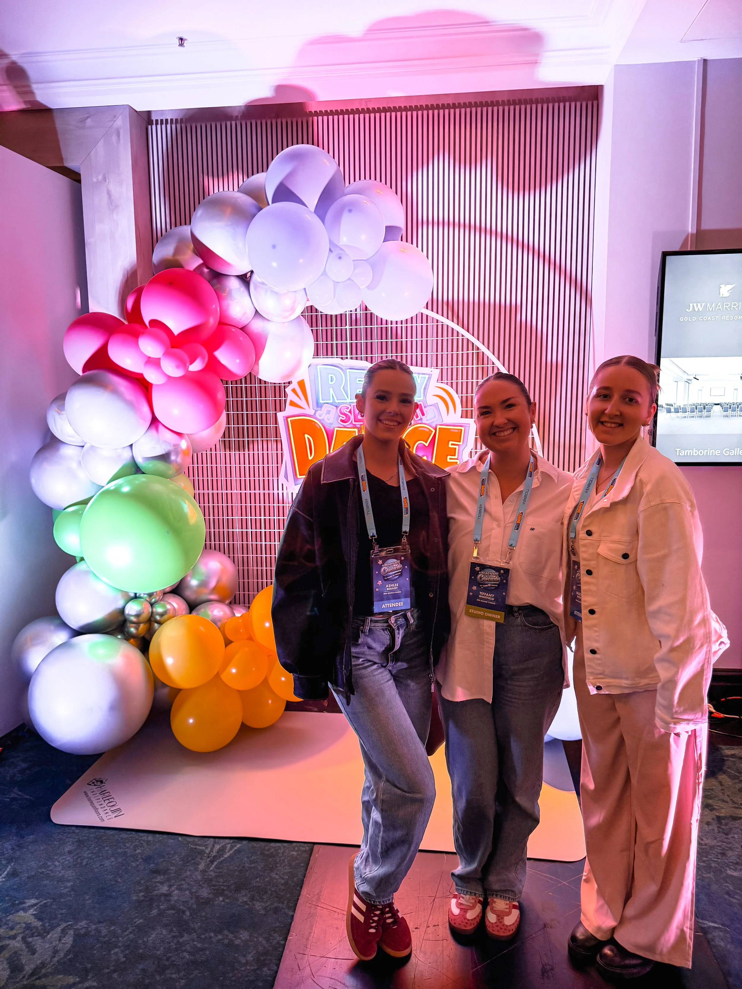 Three women standing together at an event with colorful balloon decorations and a sign that says 'RELAY SING DANCE' in the background.