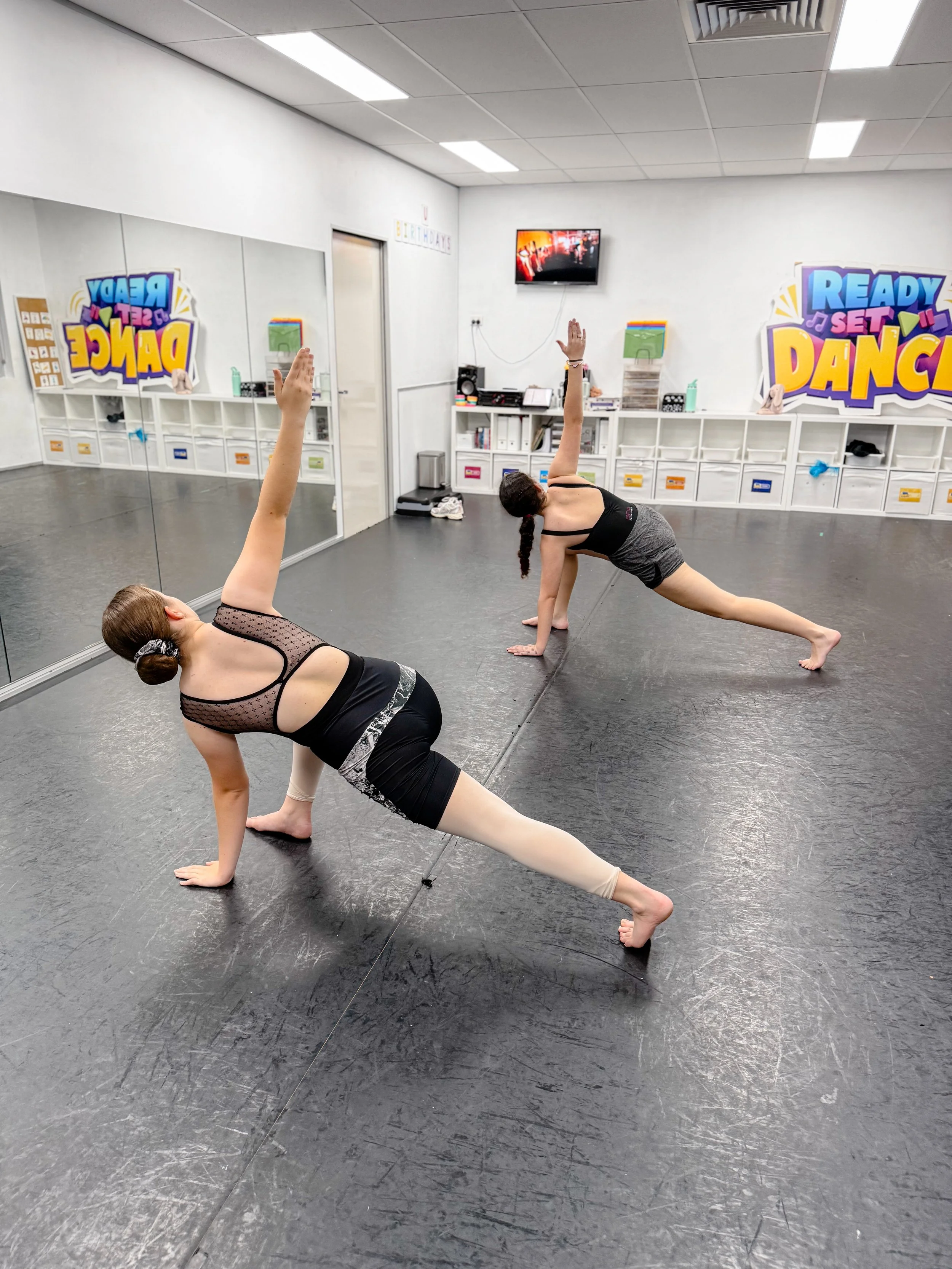 Two girls practicing yoga poses in a dance studio with a mirror, a TV on the wall, and colorful signs that say 'Ready Set Dance' and 'Ready Set Dance'.
