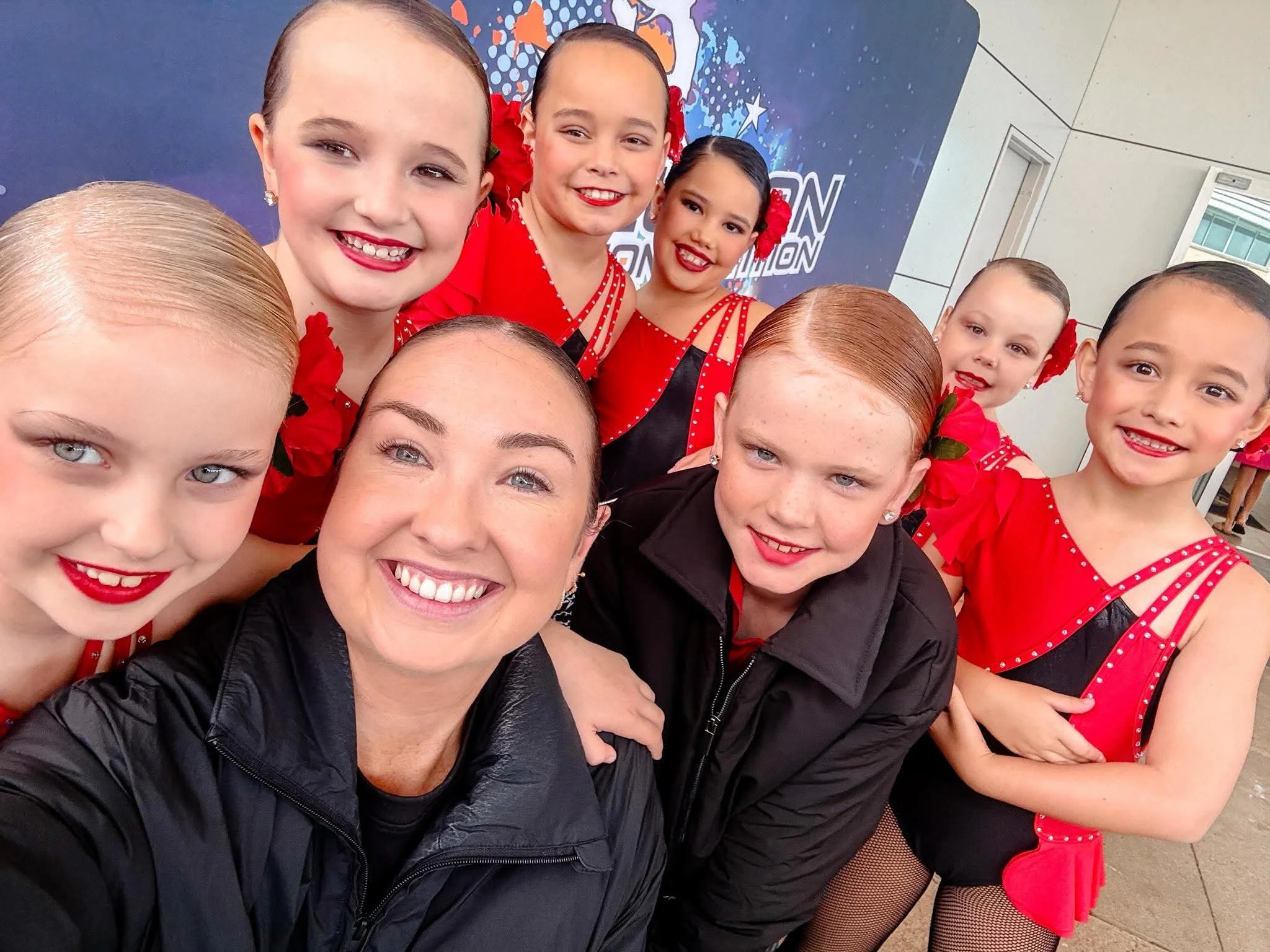 A group of seven young girls in red and black dance costumes posing for a photo, with some smiling at the camera.
