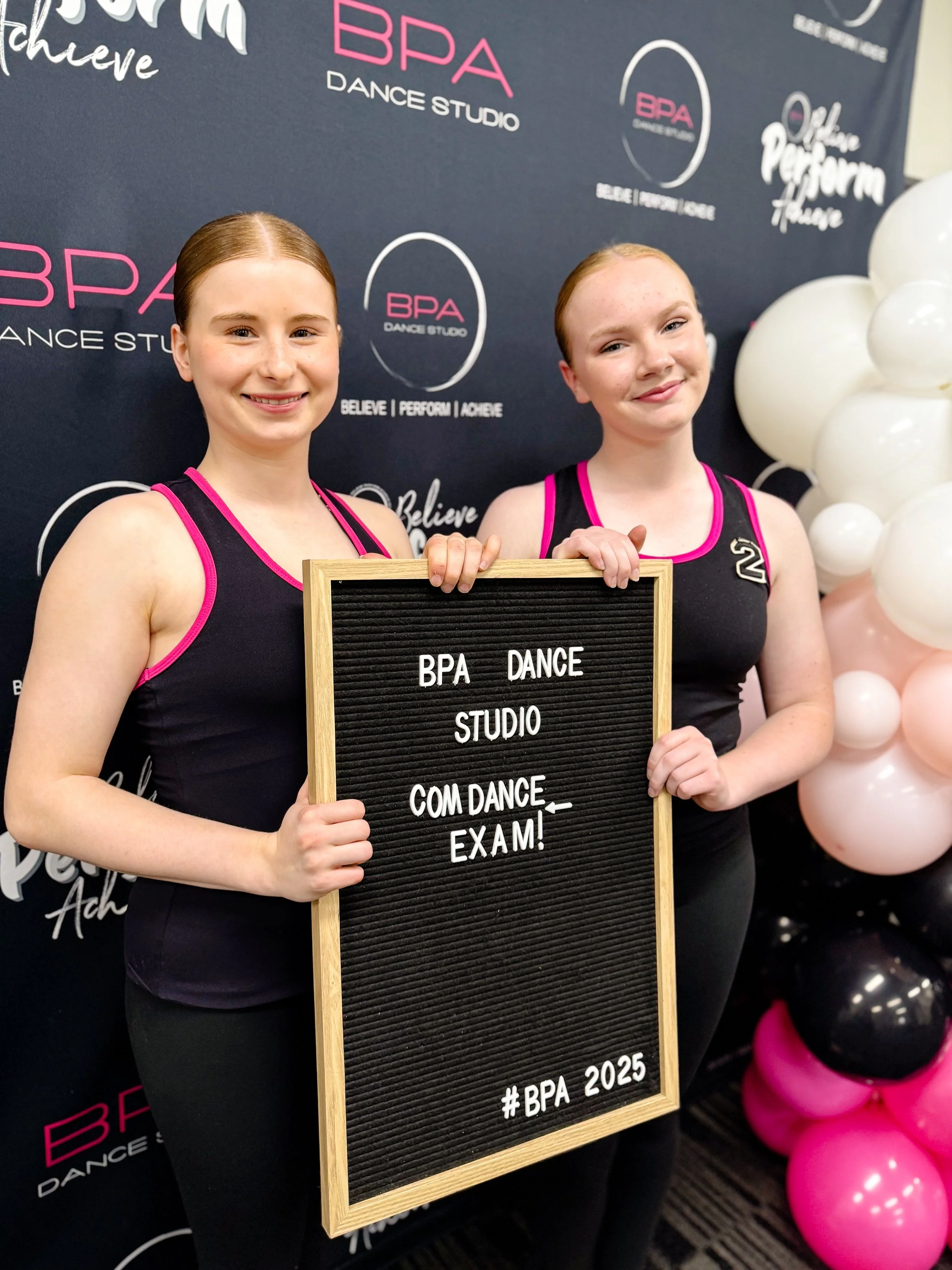 Two young women in black dance outfits with pink trim holding a black letter board at BPA Dance Studio, celebrating their exam success surrounded by pink, white, and black balloons, with the studio's branding visible on the background.