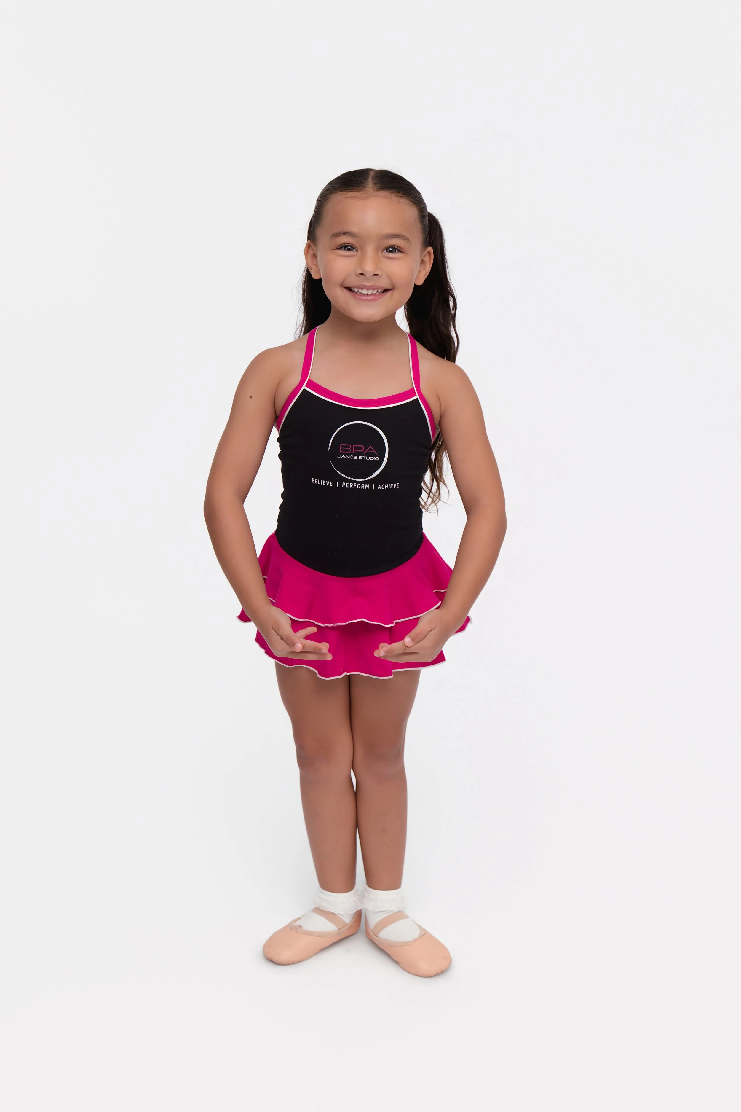 Young girl in ballet attire with pink skirt, black top featuring a dance studio logo, standing in a ballet pose with hands on her hips, smiling, against a plain white background.