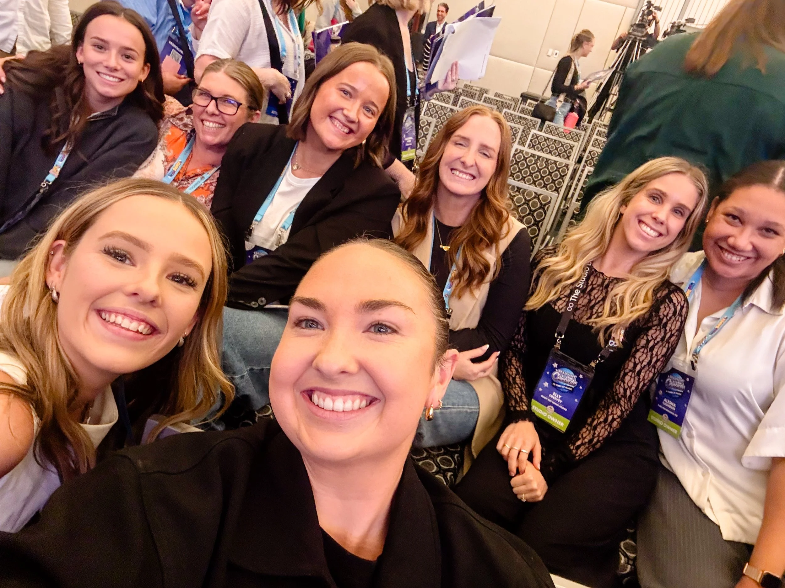 Group of women smiling and taking a selfie at a conference or event, wearing badge lanyards.
