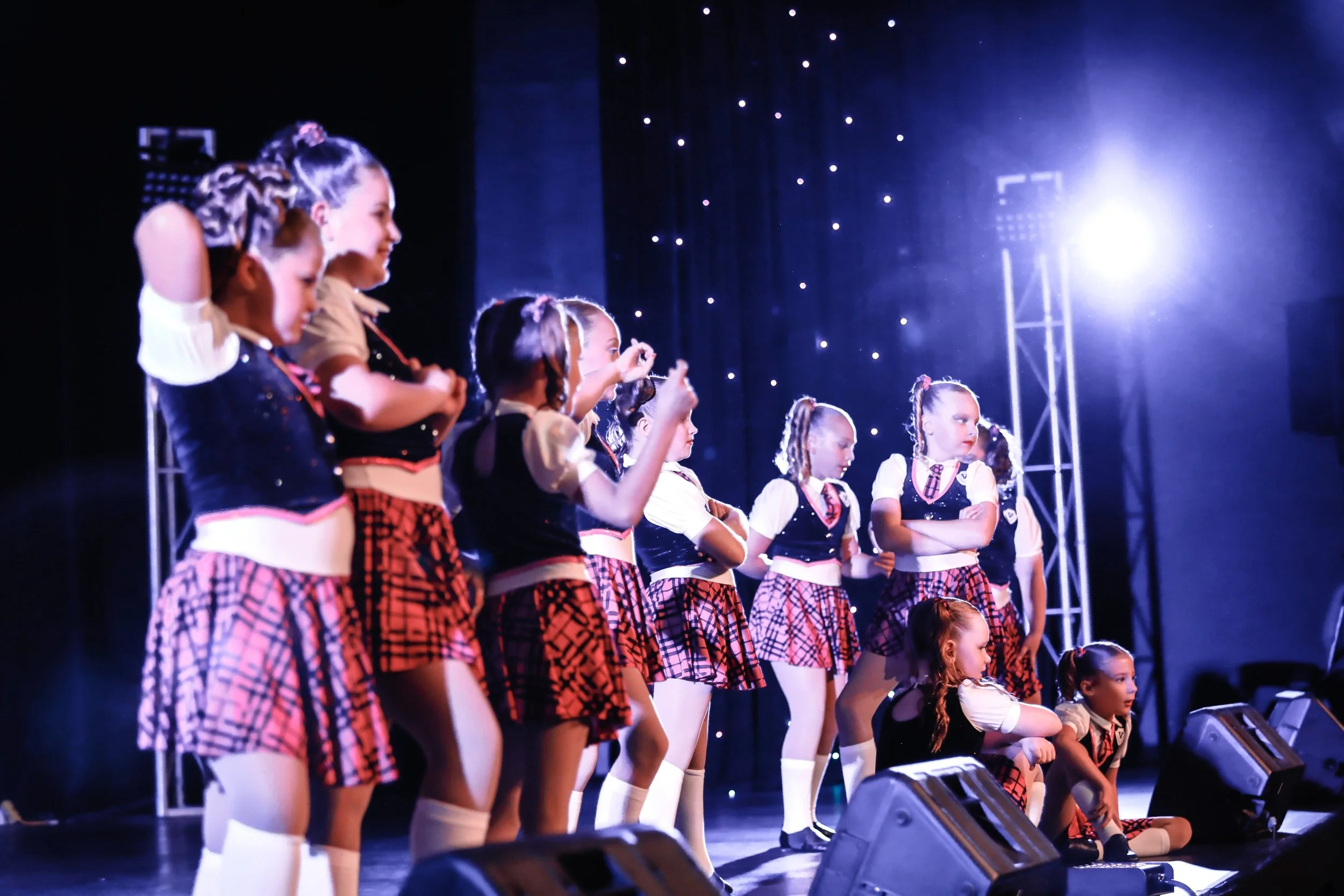 Group of young girls performing on stage, wearing matching black and red plaid costumes with white knee-high socks, under stage lighting with a black starry backdrop.