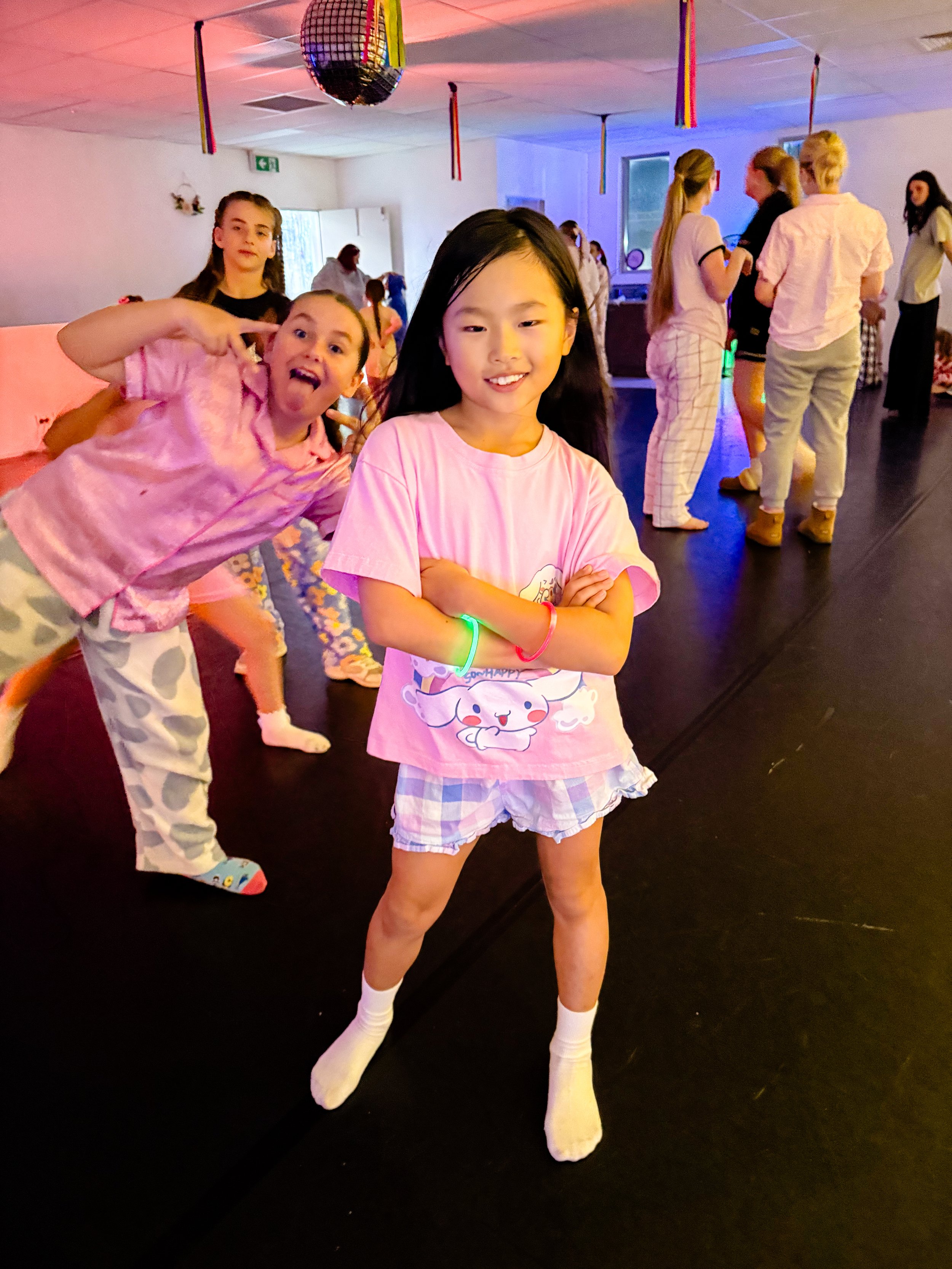 Children at a party or dance event, one girl in the foreground with arms crossed smiling, others in pajamas and casual clothes in the background, colorful decorations hanging from the ceiling, a disco ball, and party lighting.