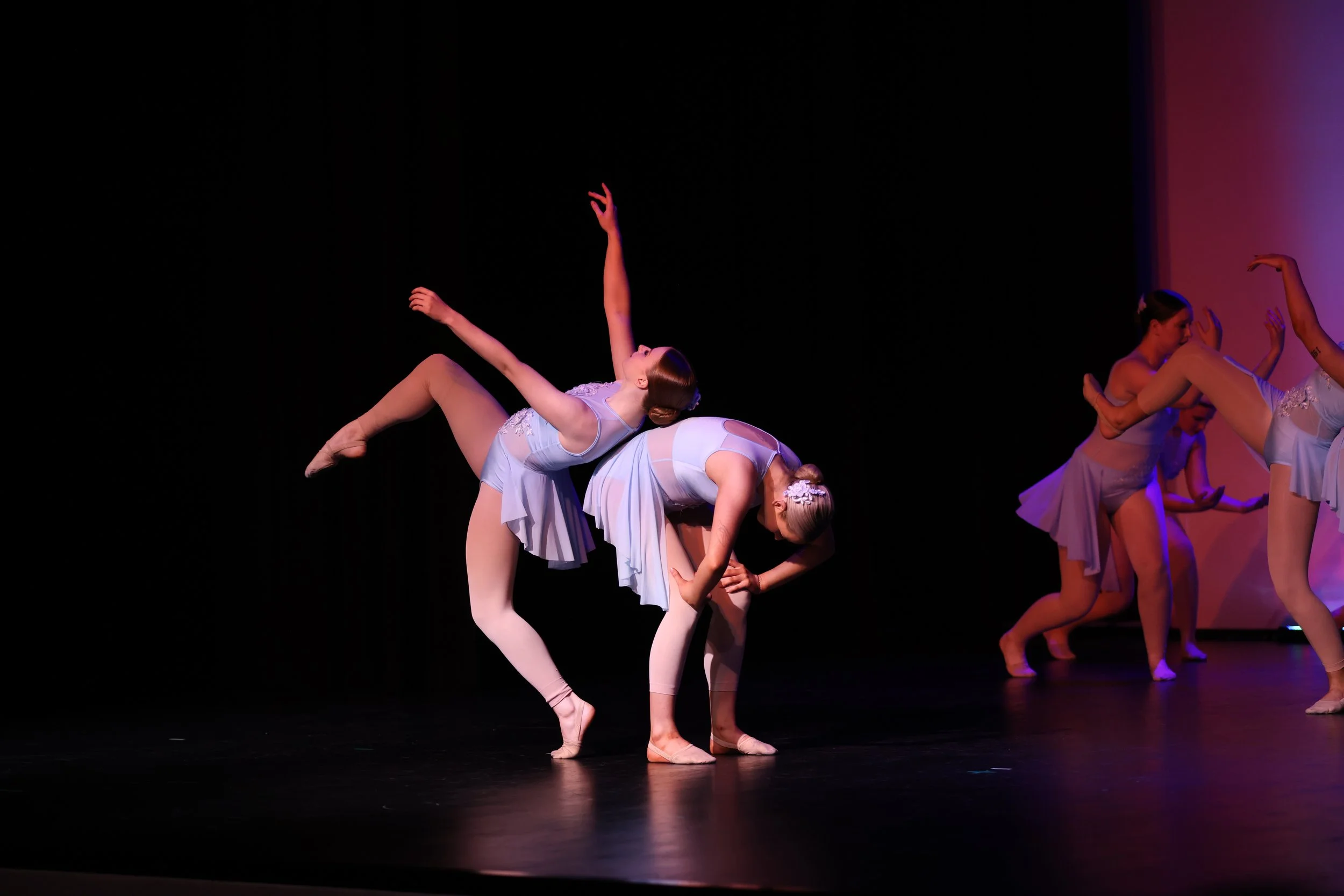 Ballet dancers on stage, with some bending forward and others raising arms, dressed in light blue costumes, under stage lights.