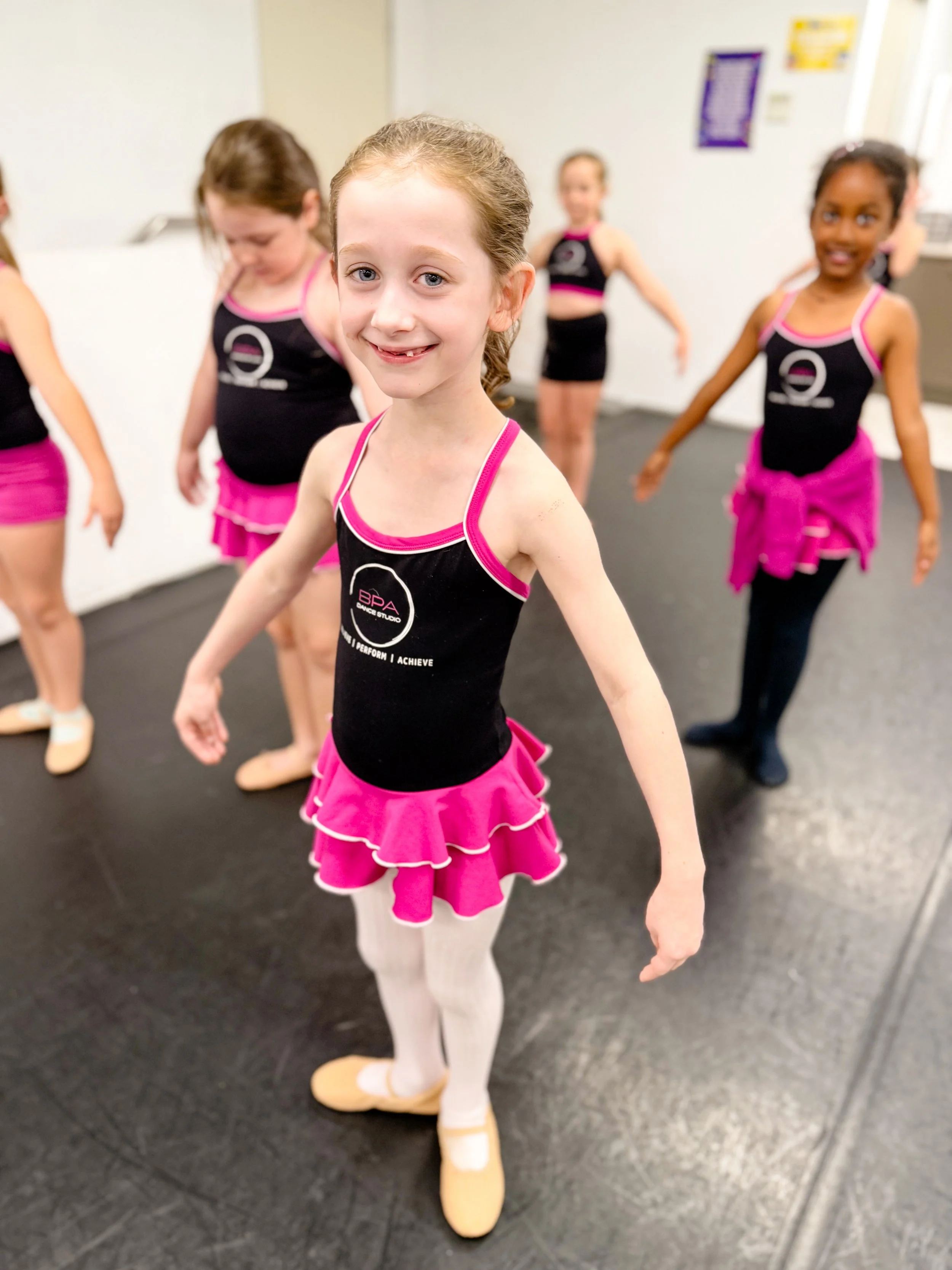 A young girl in a black and pink ballet costume standing in front with her arms out, smiling, and other girls in similar costumes behind her in a dance studio.