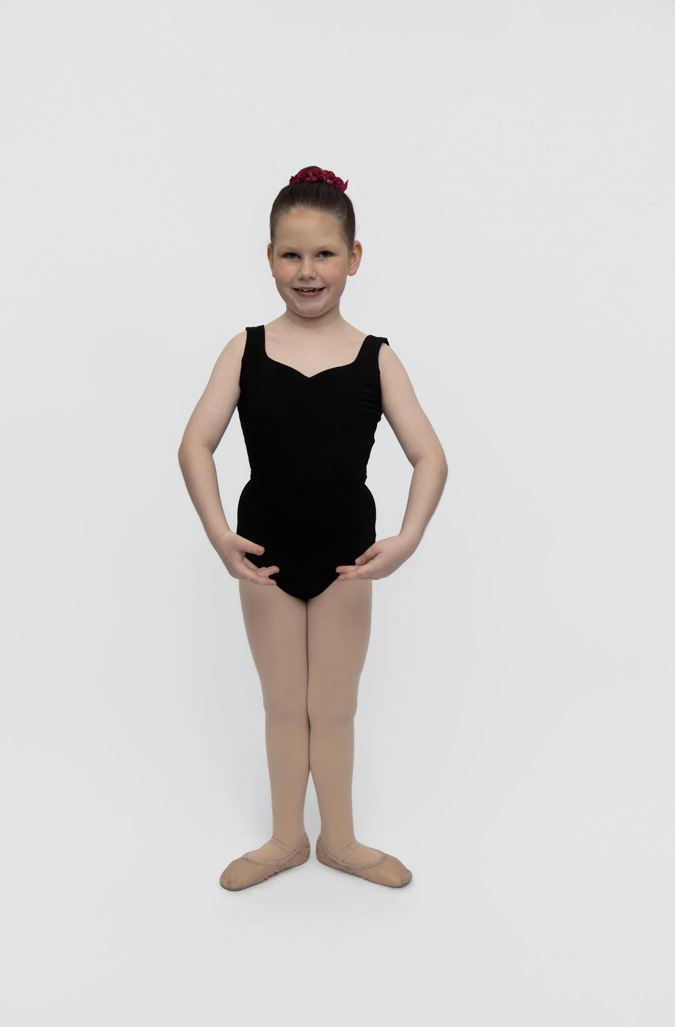 Young girl in a black ballet leotard, beige tights, ballet slippers, and a red hair accessory, standing in a ballet pose against a plain white background.