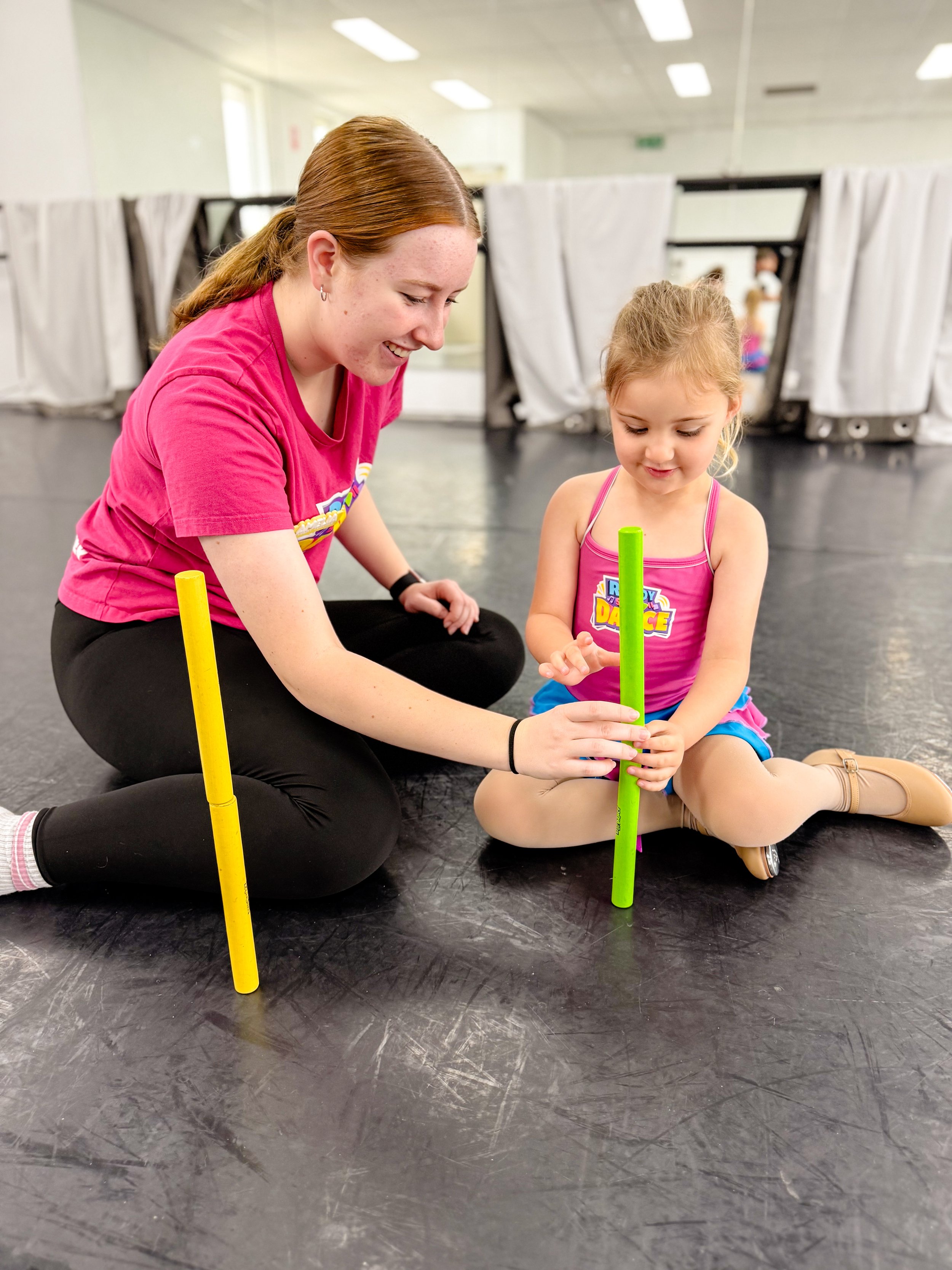 A young girl and an adult woman sitting on the floor of a dance studio, playing with foam sticks.