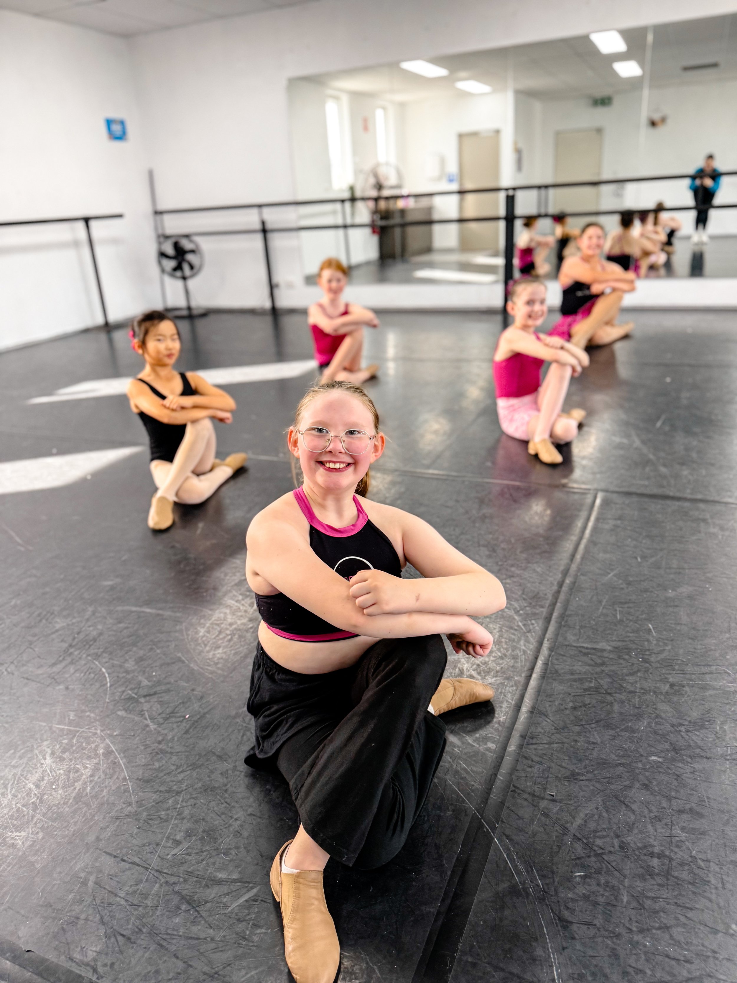 A group of young ballet dancers, mostly girls, sitting on the floor of a dance studio, with one girl in the foreground smiling at the camera. The studio has black flooring, a ballet barre, and a large mirror.