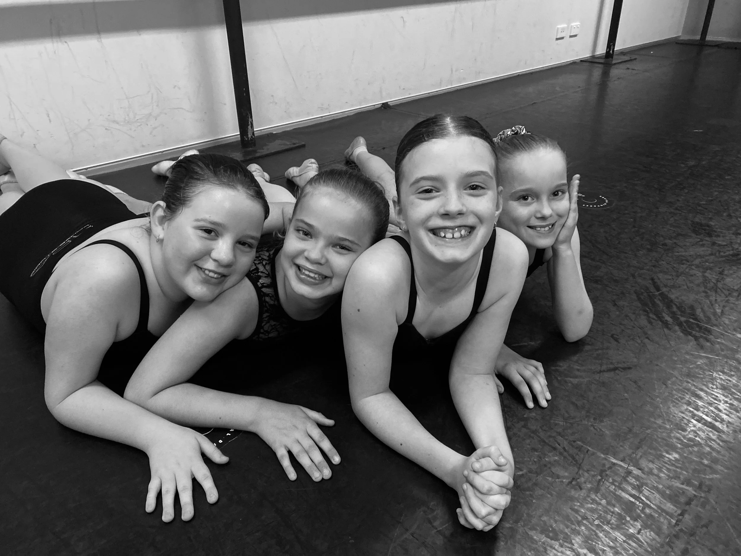 Four young girls lying on a dance studio floor, smiling at the camera.
