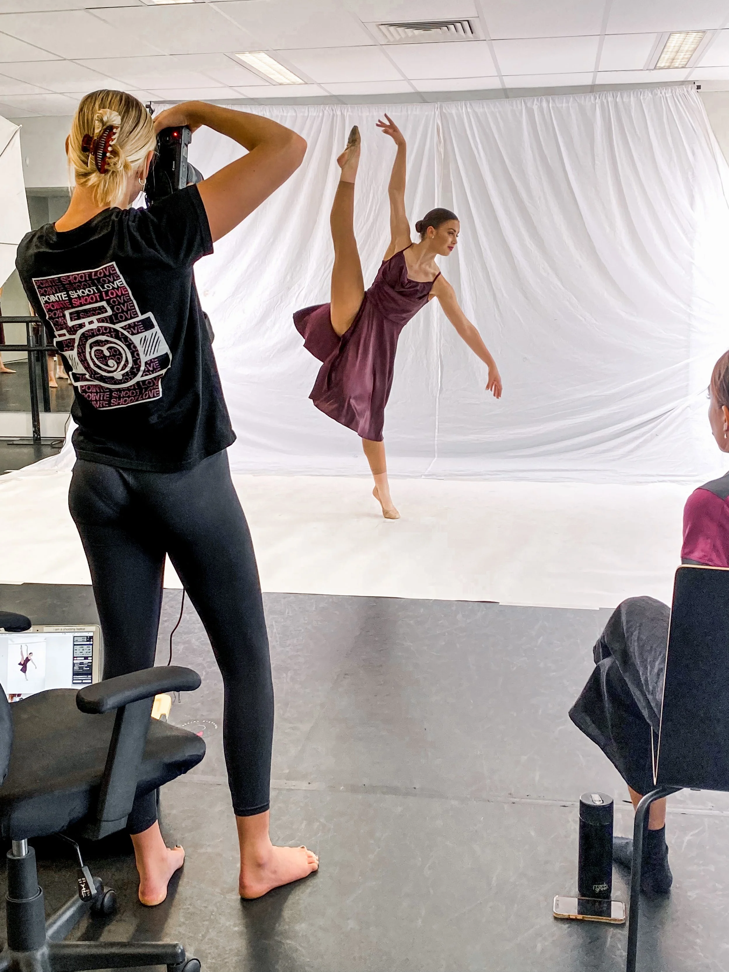 A ballet dancer in a purple dress practices a high leg extension in front of a white backdrop, while being filmed by a dancer with a camera and two seated individuals observe.
