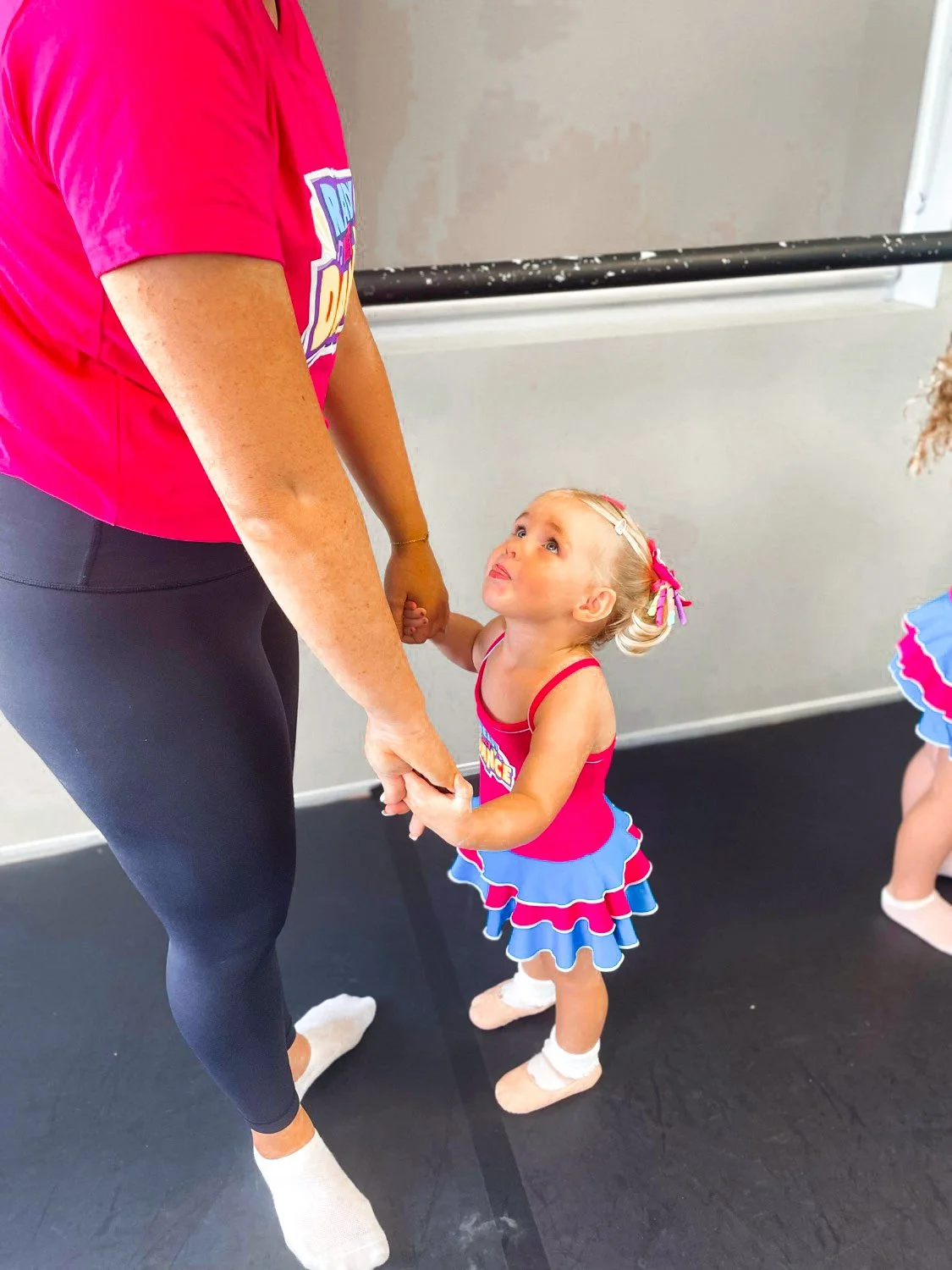 A young girl in a pink and blue cheerleading outfit holding hands with an adult woman in black leggings and a pink shirt, in a dance studio.