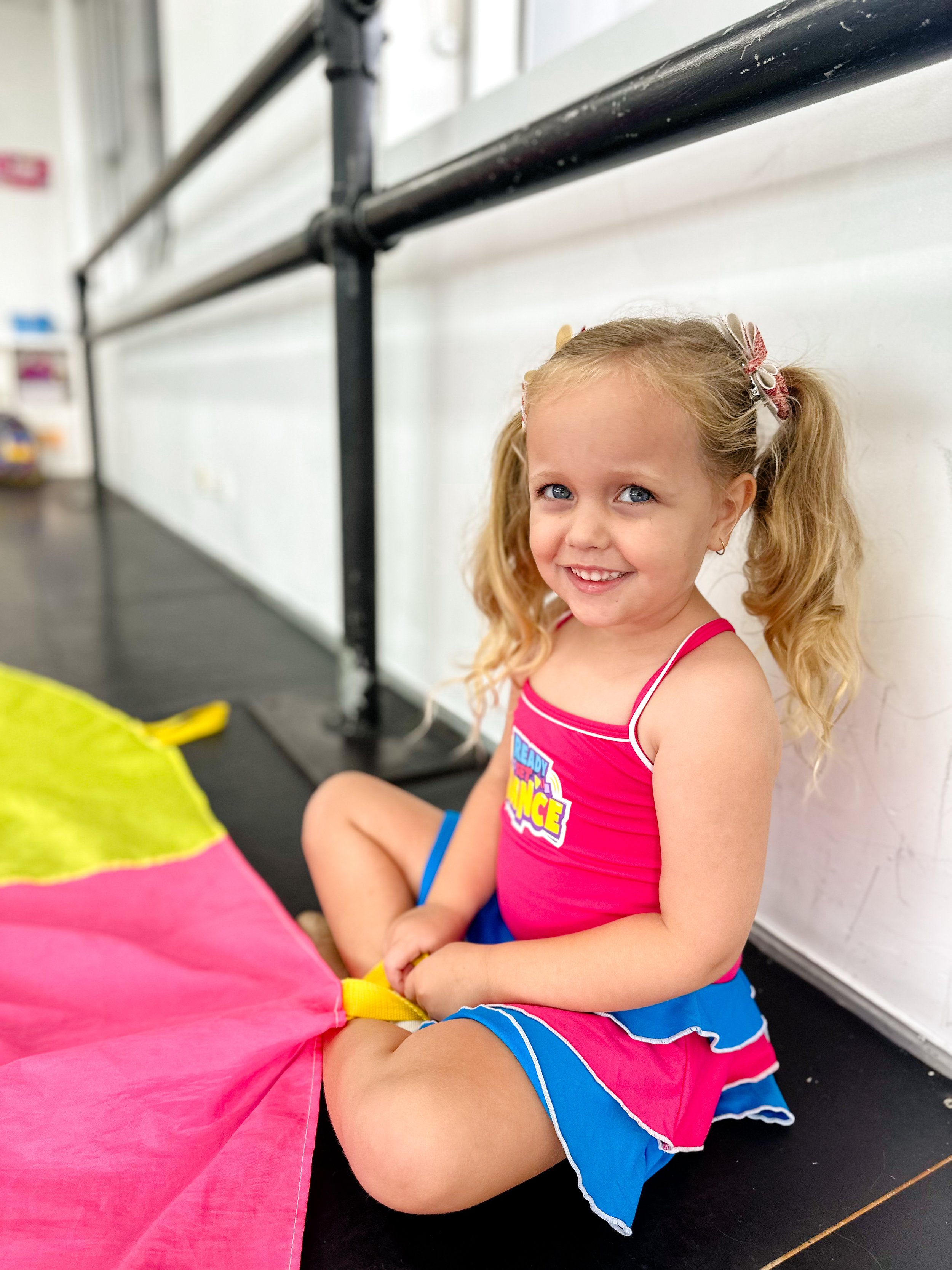 A young girl with blonde hair and blue eyes sitting on the floor, smiling, wearing a pink, blue, and white cheerleading outfit, holding a pink and yellow parachute.