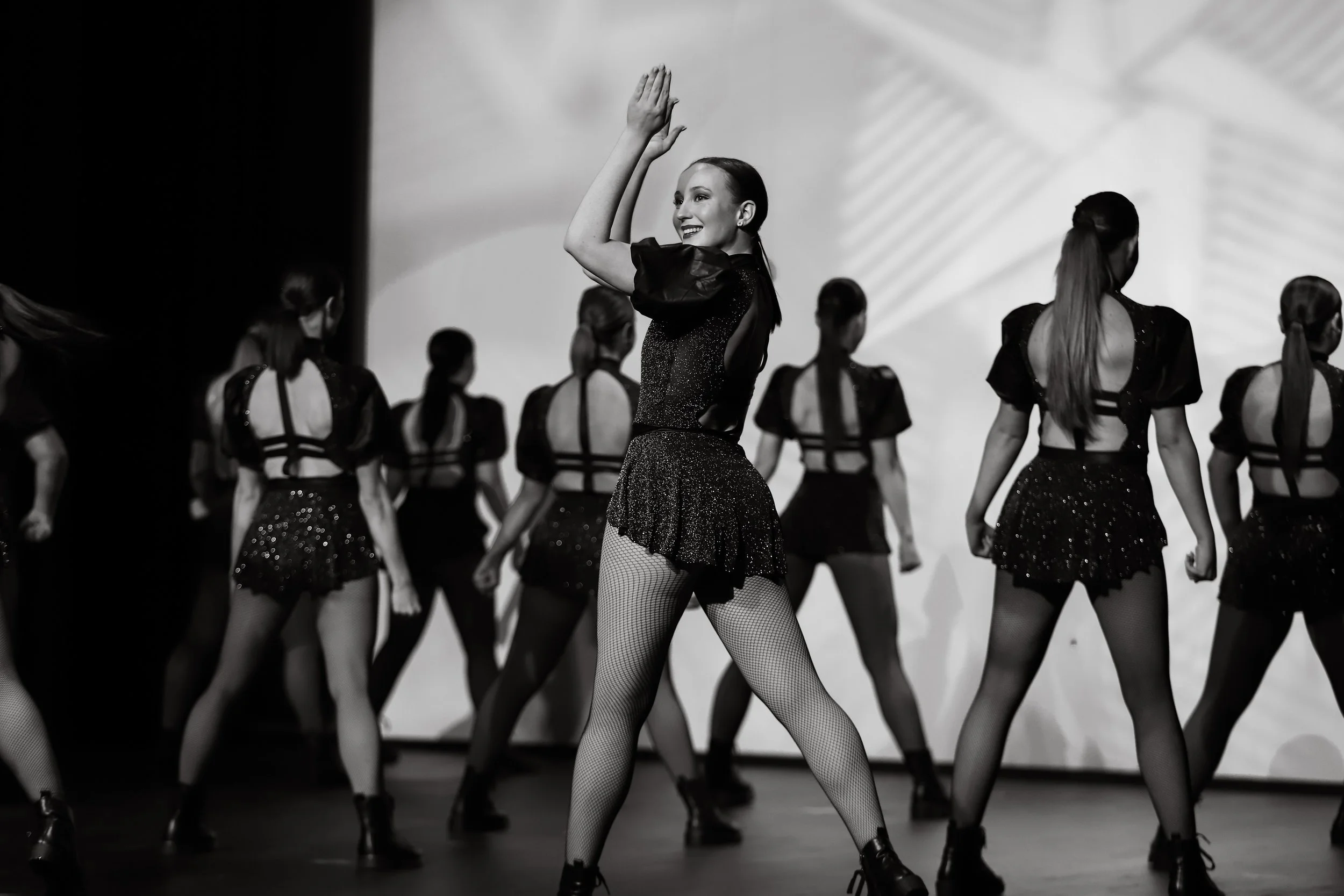 A dance performance featuring women in black costumes on stage. One woman is front and center, smiling and posing with her hands raised, while the others are in the background, facing away or side-facing.