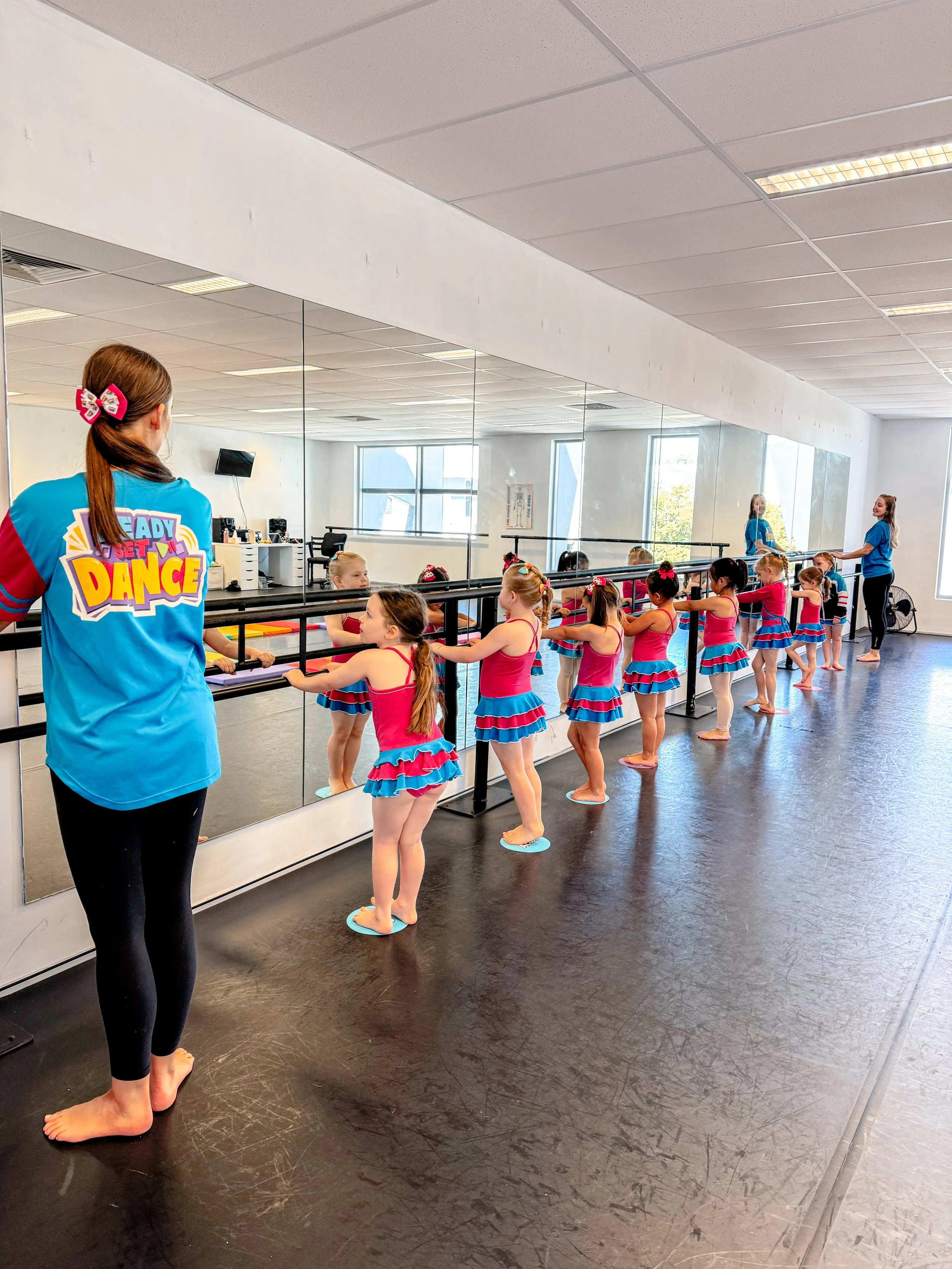 Young ballet students in pink and blue tutus practicing at a ballet barre in a dance studio. An instructor is supervising.